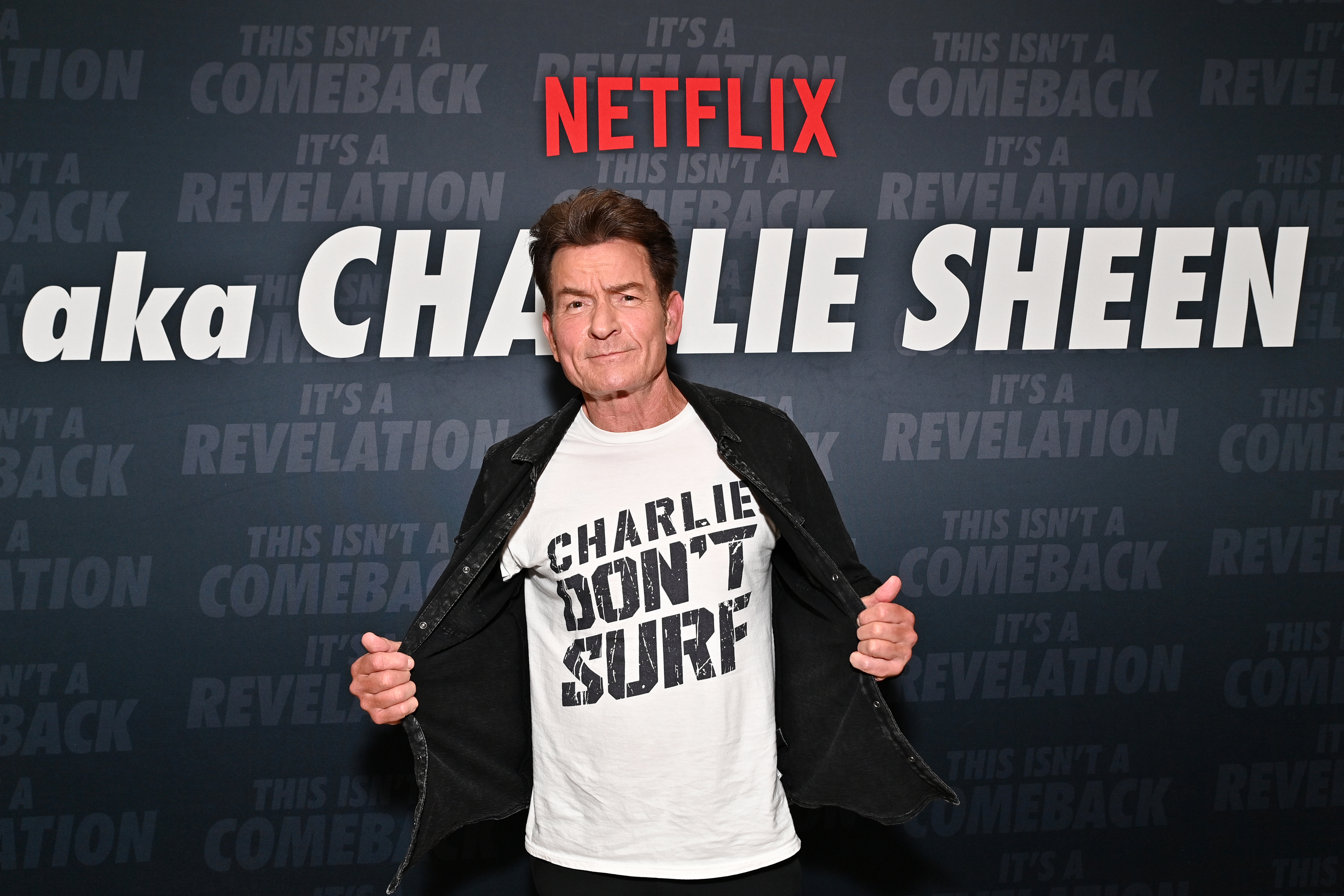 Charlie Sheen opens his black jacket to show a white 'Charlie Don't Surf' t-shirt in front of a Netflix 'aka Charlie Sheen' backdrop.