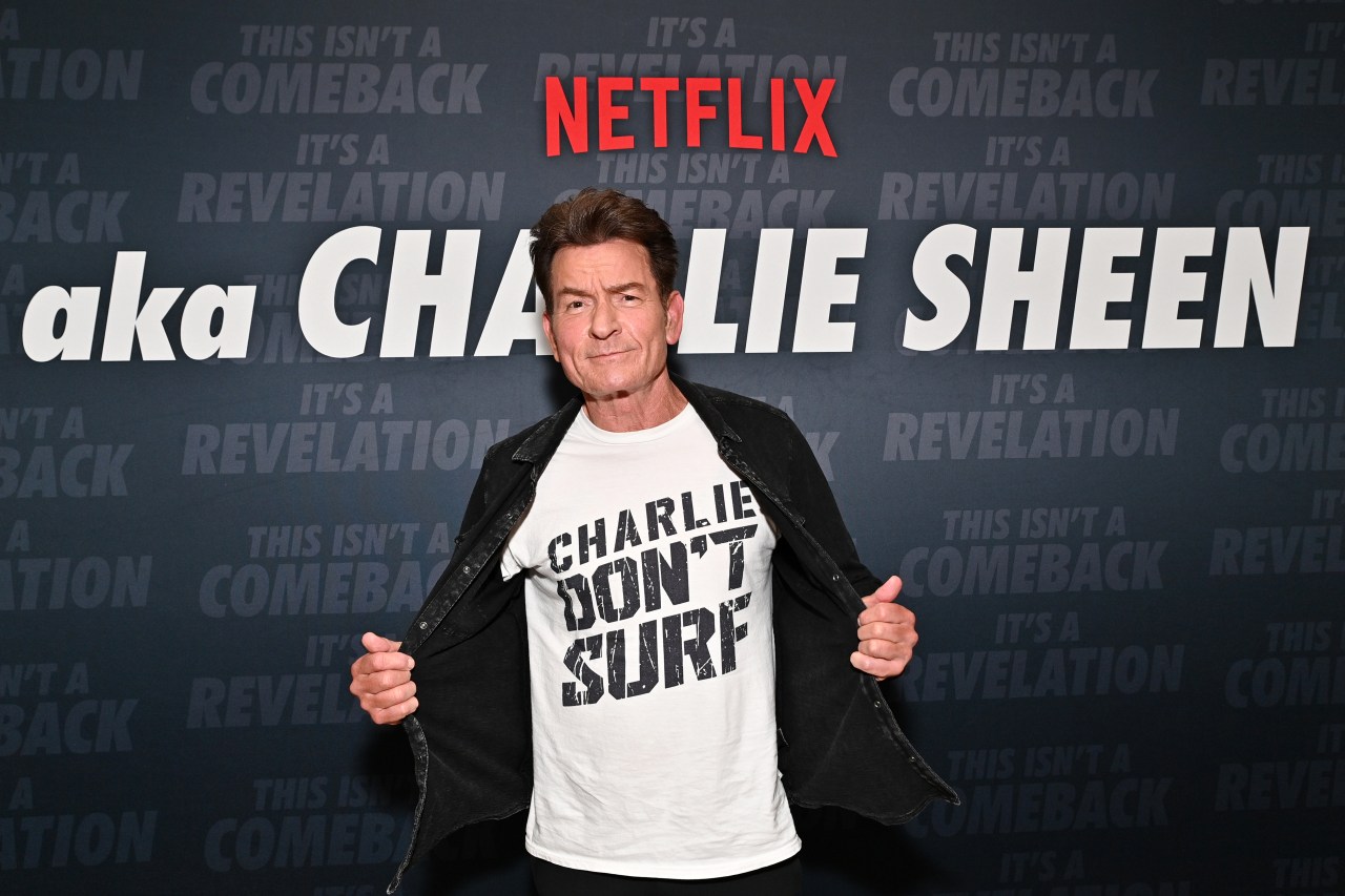 Charlie Sheen opens his black jacket to show a white 'Charlie Don't Surf' t-shirt in front of a Netflix 'aka Charlie Sheen' backdrop.