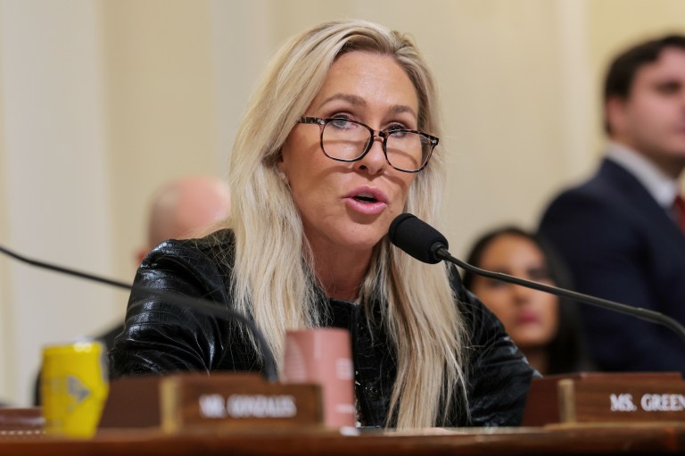 Rep. Marjorie Taylor Greene speaks into a microphone at a congressional hearing, wearing glasses and a black jacket, with a nameplate reading 'Ms. Green' visible on the desk.