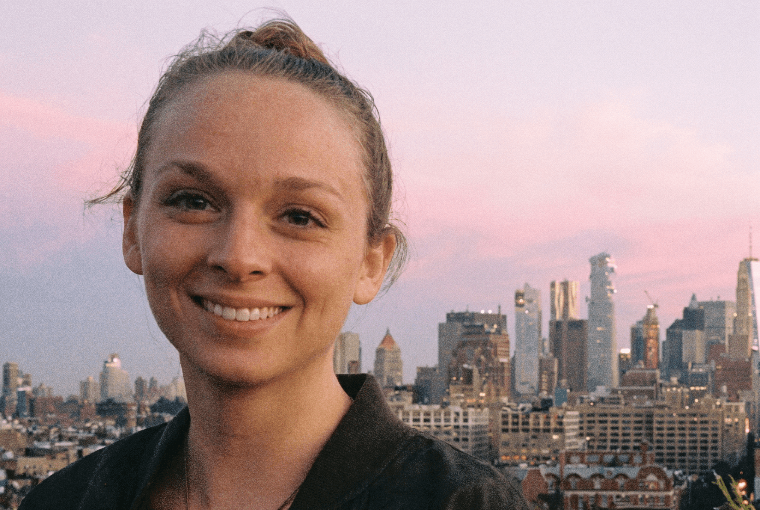 A smiling young woman with hair pulled back poses outdoors with a city skyline and pink sunset sky in the background.