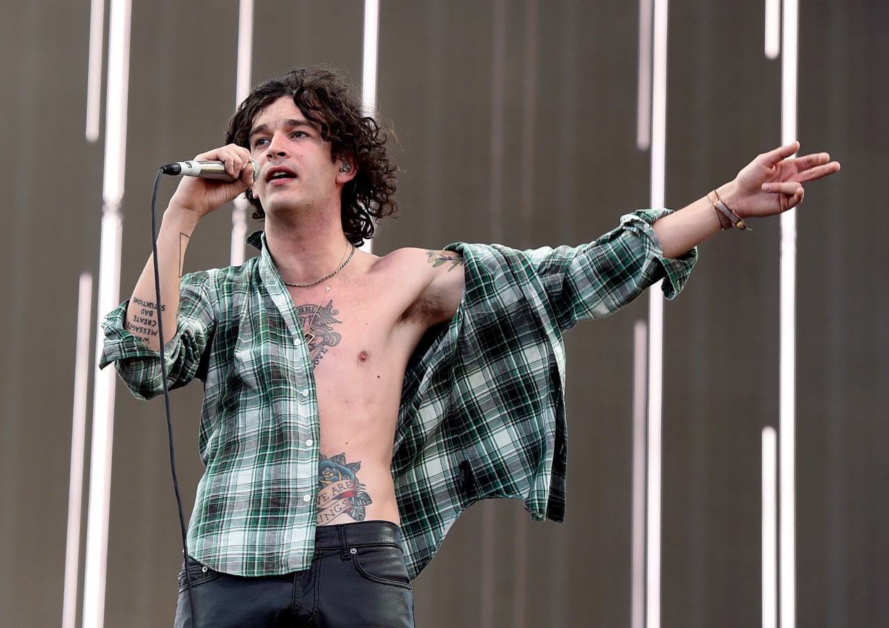 Curly-haired male singer in an open green plaid shirt performing with a microphone on a festival stage, one arm outstretched.