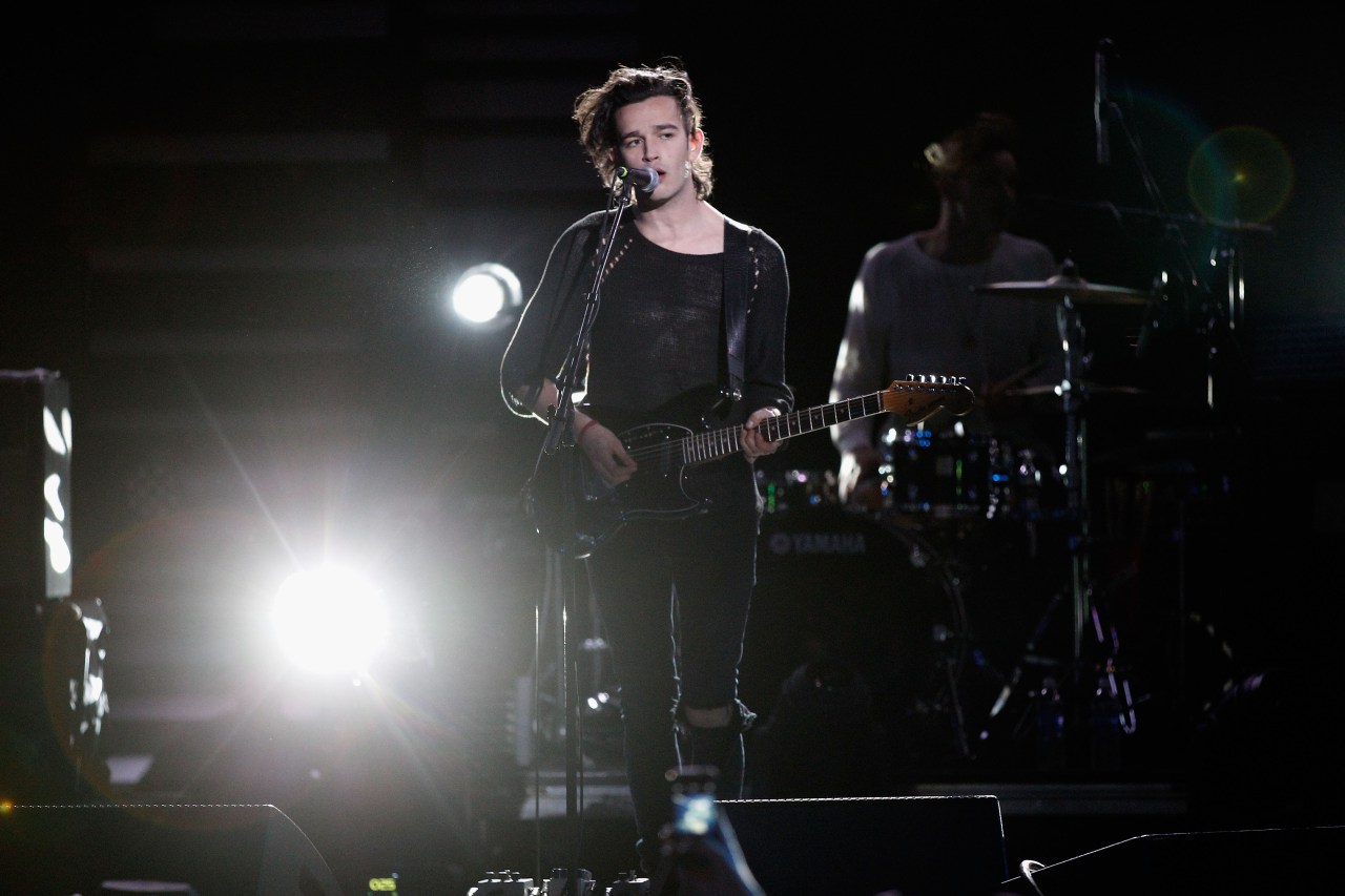 Young male musician playing a black electric guitar and singing into a microphone on a darkened stage, with a drummer visible behind him.