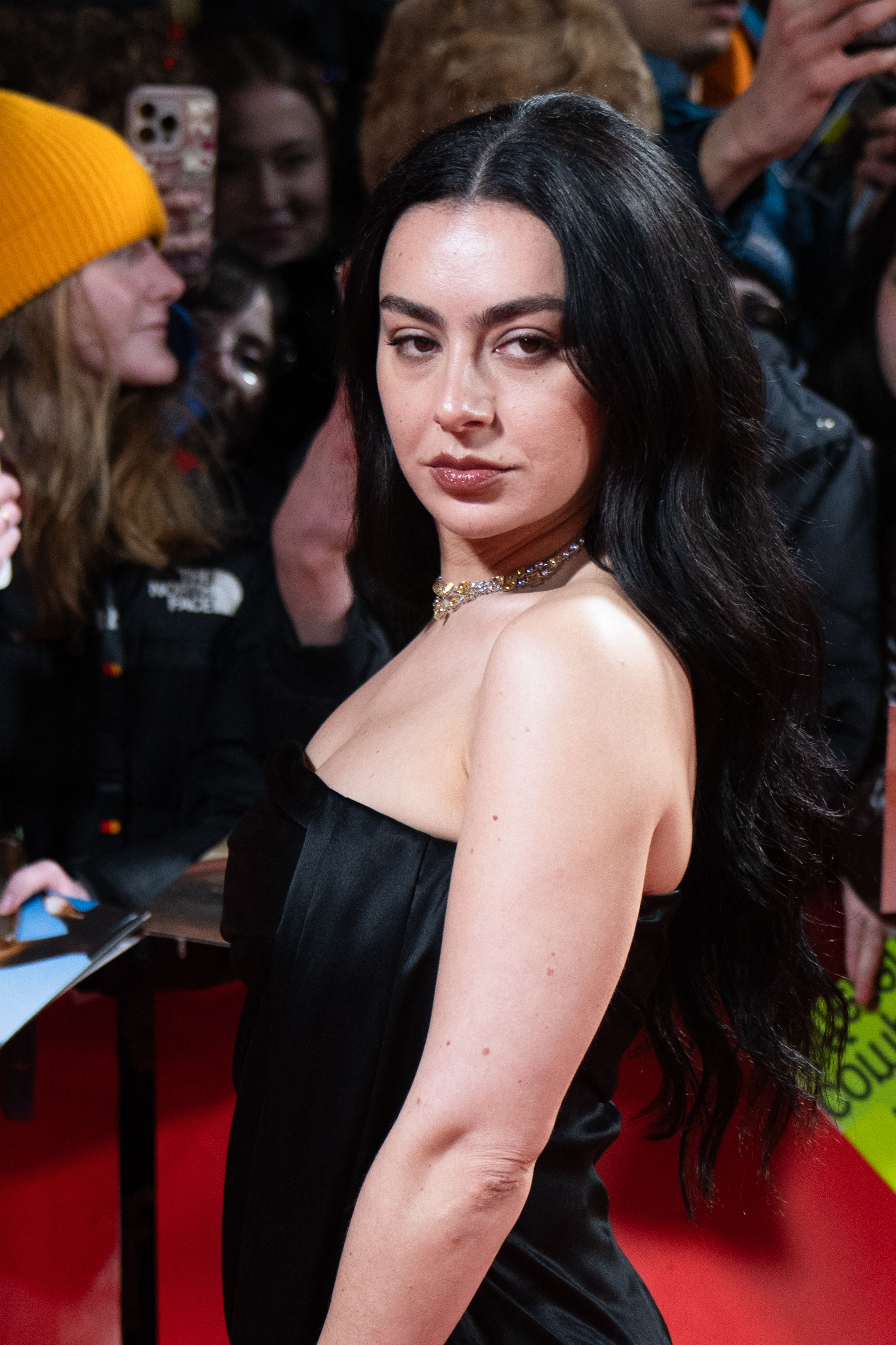 Charli XCX in a strapless black dress with a jeweled choker on the red carpet at the Berlinale Film Festival, fans visible in the background.