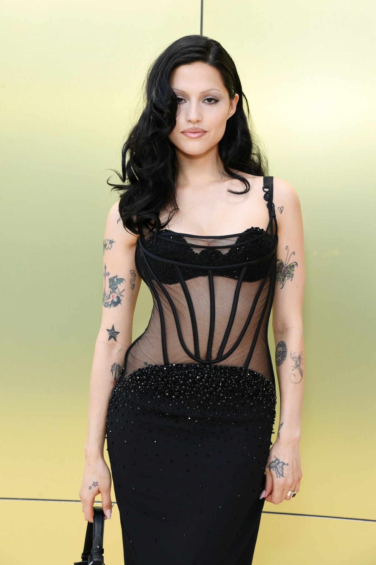 Dark-haired tattooed woman in a sheer black corset gown with beaded skirt, posing against a gold wall.