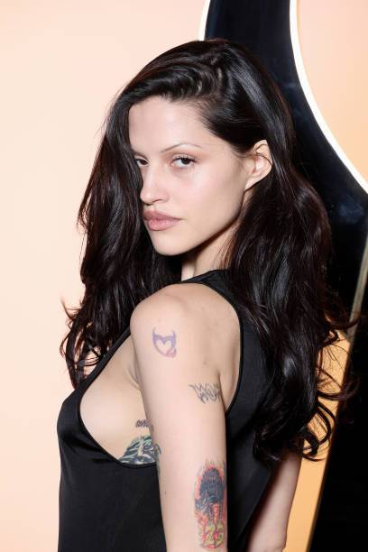 Dark-haired woman in a black sleeveless dress with multiple arm tattoos, posing in front of a dark curved backdrop.
