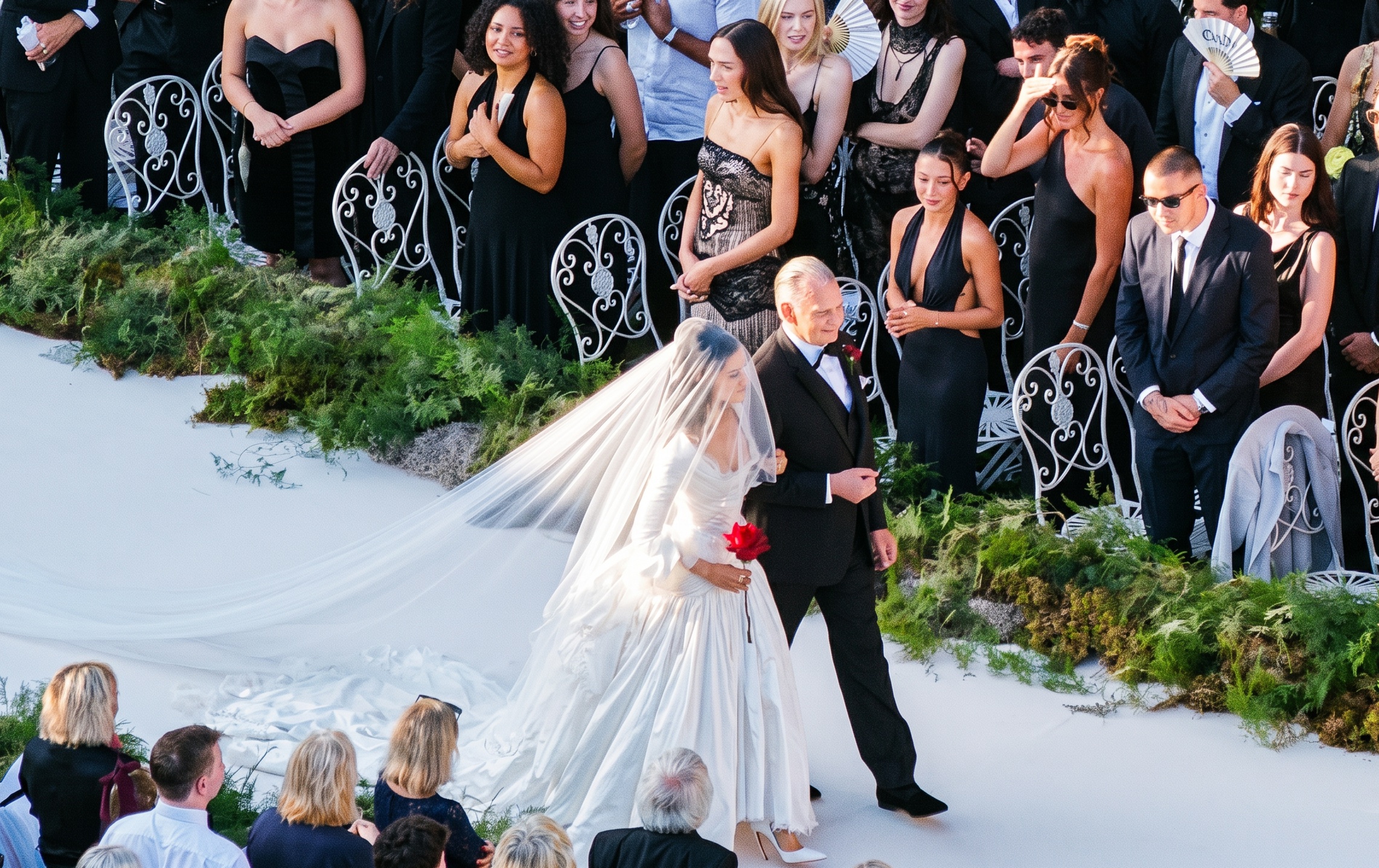 Bride in long white gown and veil holding a red rose, escorted down a white aisle by a man in a tuxedo, surrounded by guests in black attire.