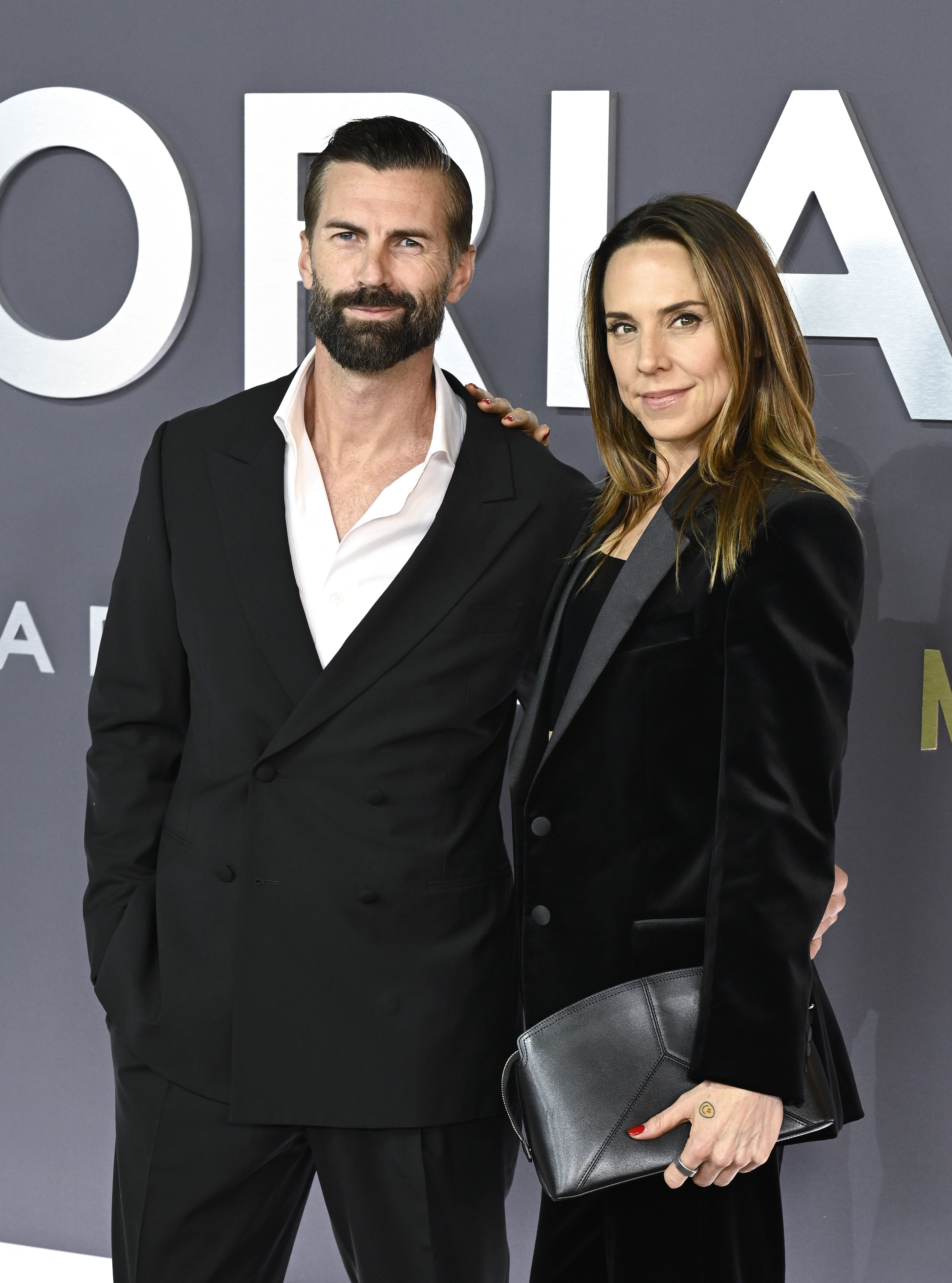 A bearded man in a black double-breasted suit and a brunette woman in a black velvet blazer holding a leather clutch pose together on a red carpet.