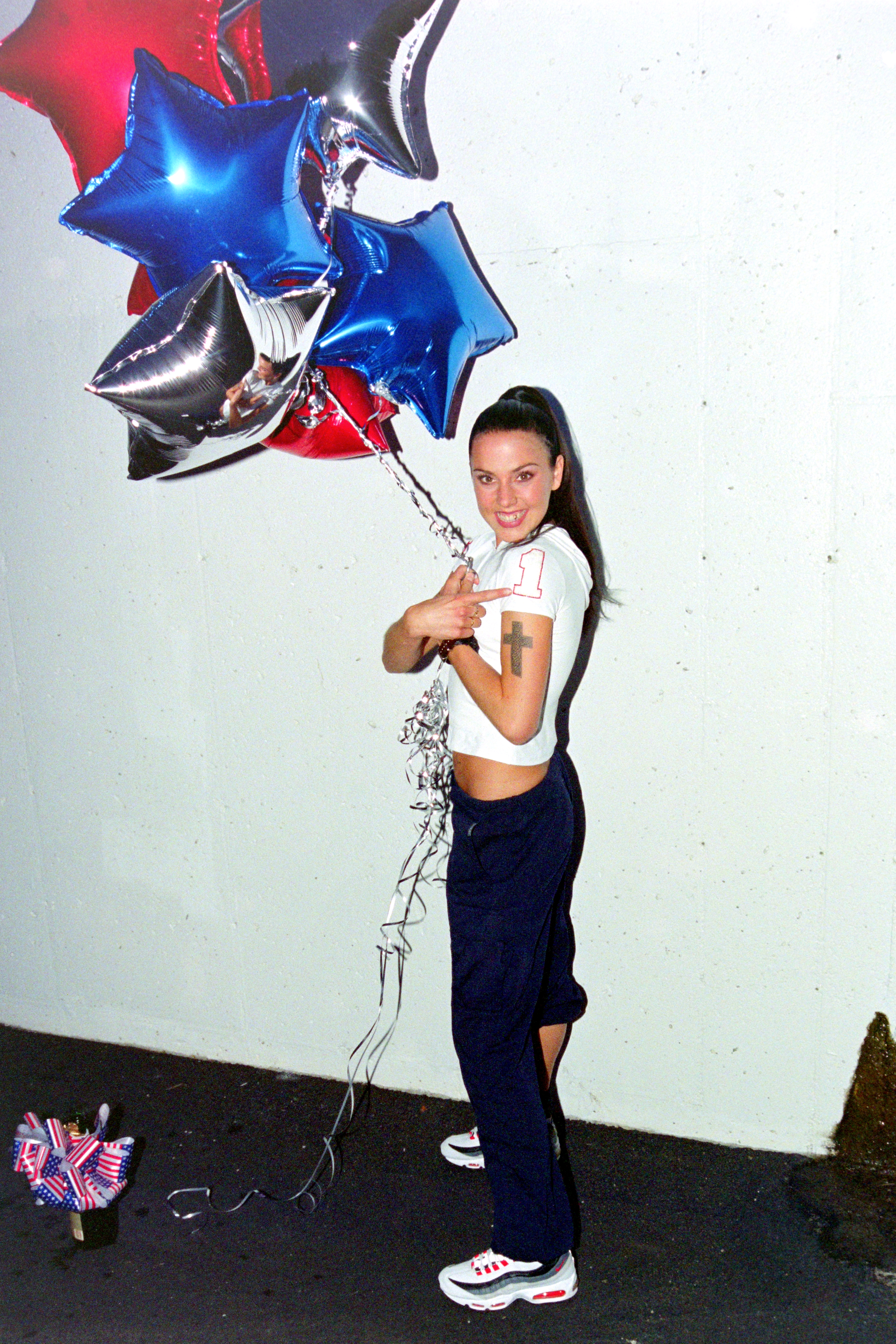 Young woman in a white crop top and navy track pants holds red, blue, and silver star-shaped foil balloons, smiling and pointing at them.