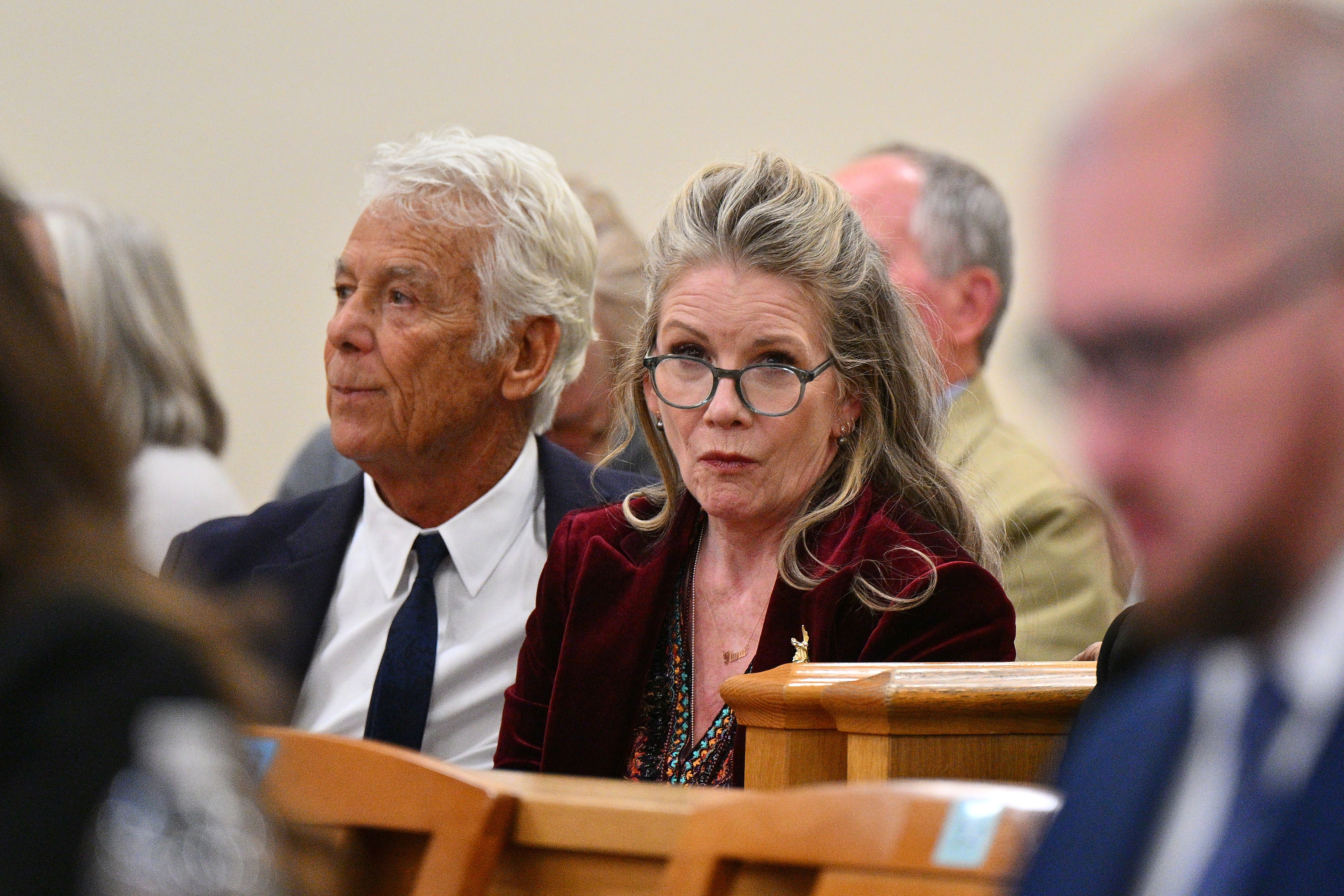 Click to open the image gallery Melissa Gilbert wearing glasses and a burgundy velvet jacket sits beside an older man in a navy suit inside a courtroom.