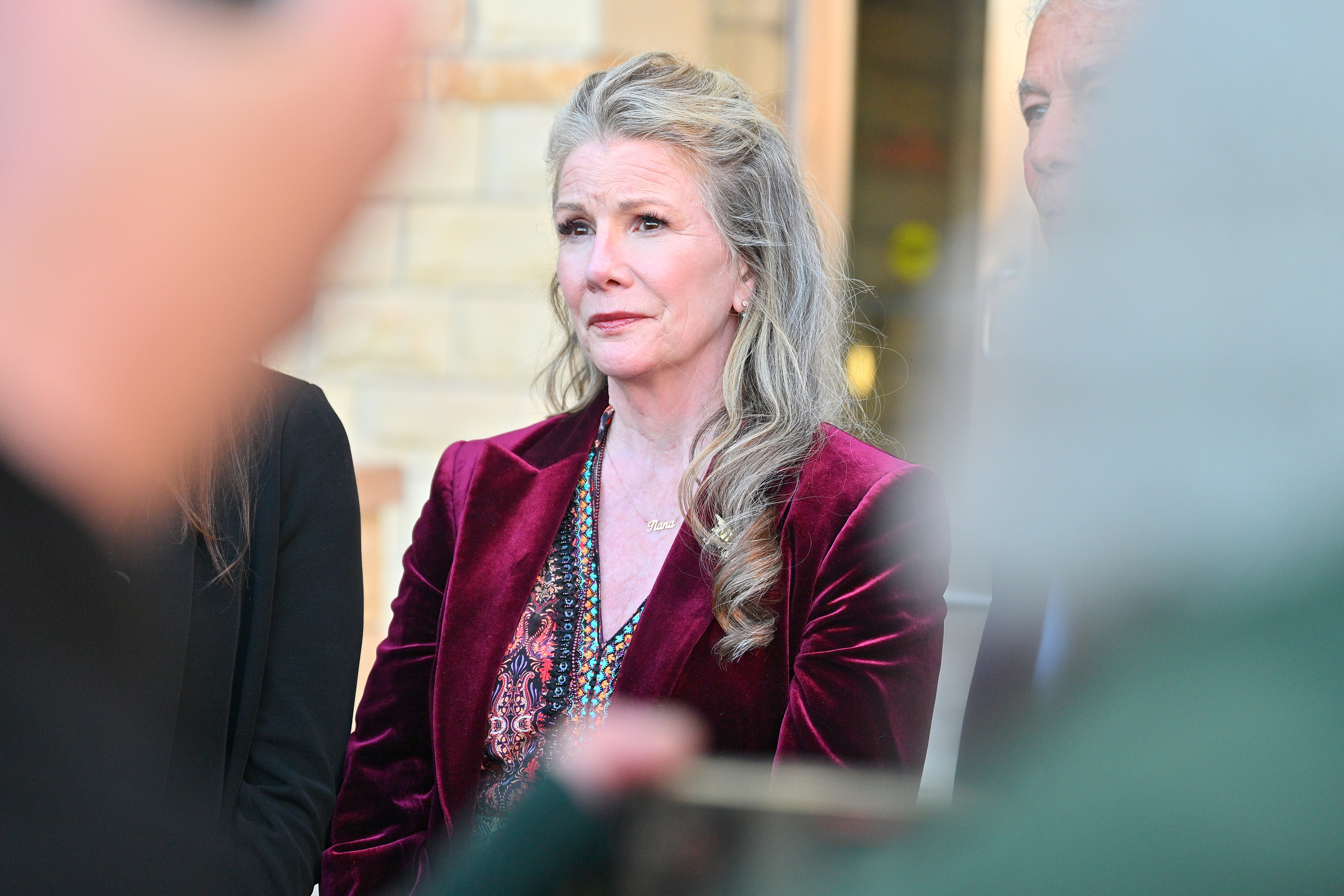 Click to open the image gallery A woman with long gray-blonde hair in a burgundy velvet blazer stands outdoors, looking ahead with a somber expression.