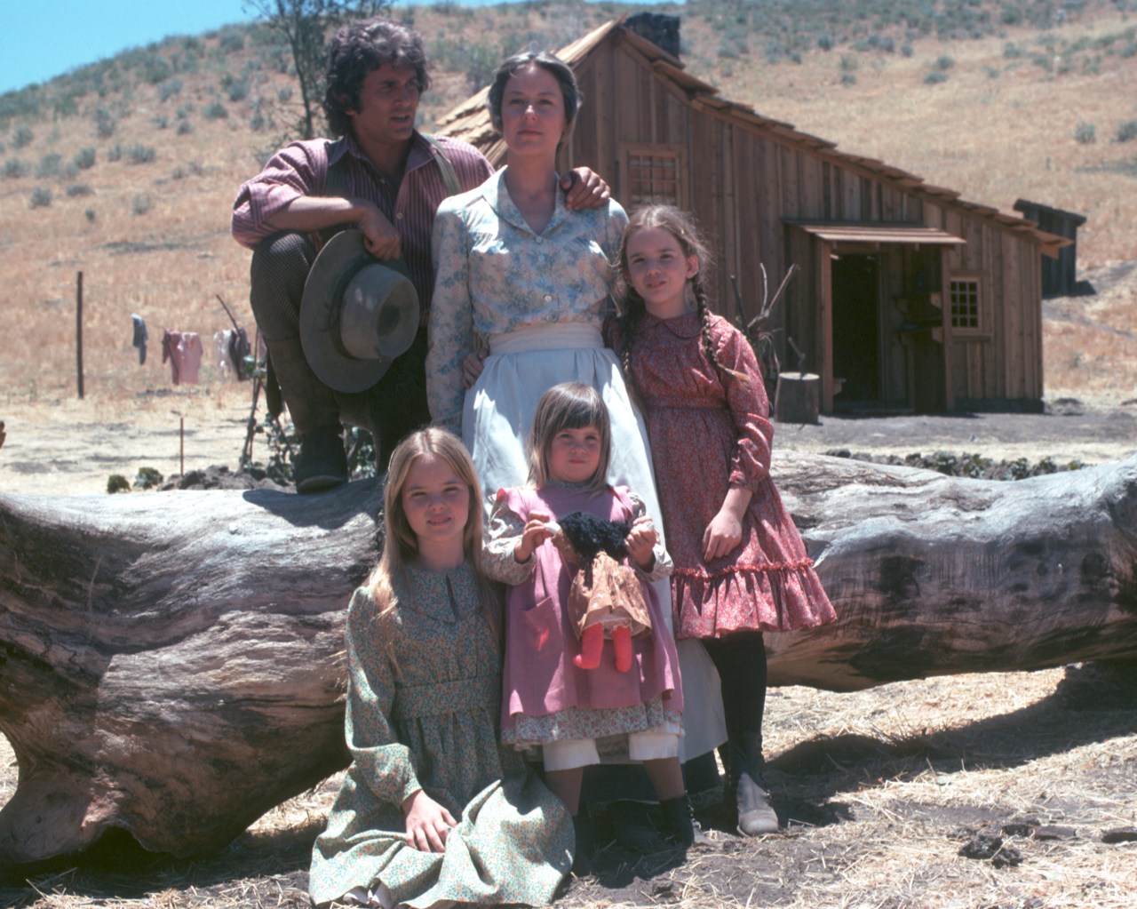 A man and woman in pioneer-era clothes pose with three young girls in front of a weathered wooden cabin on a dry, open landscape.