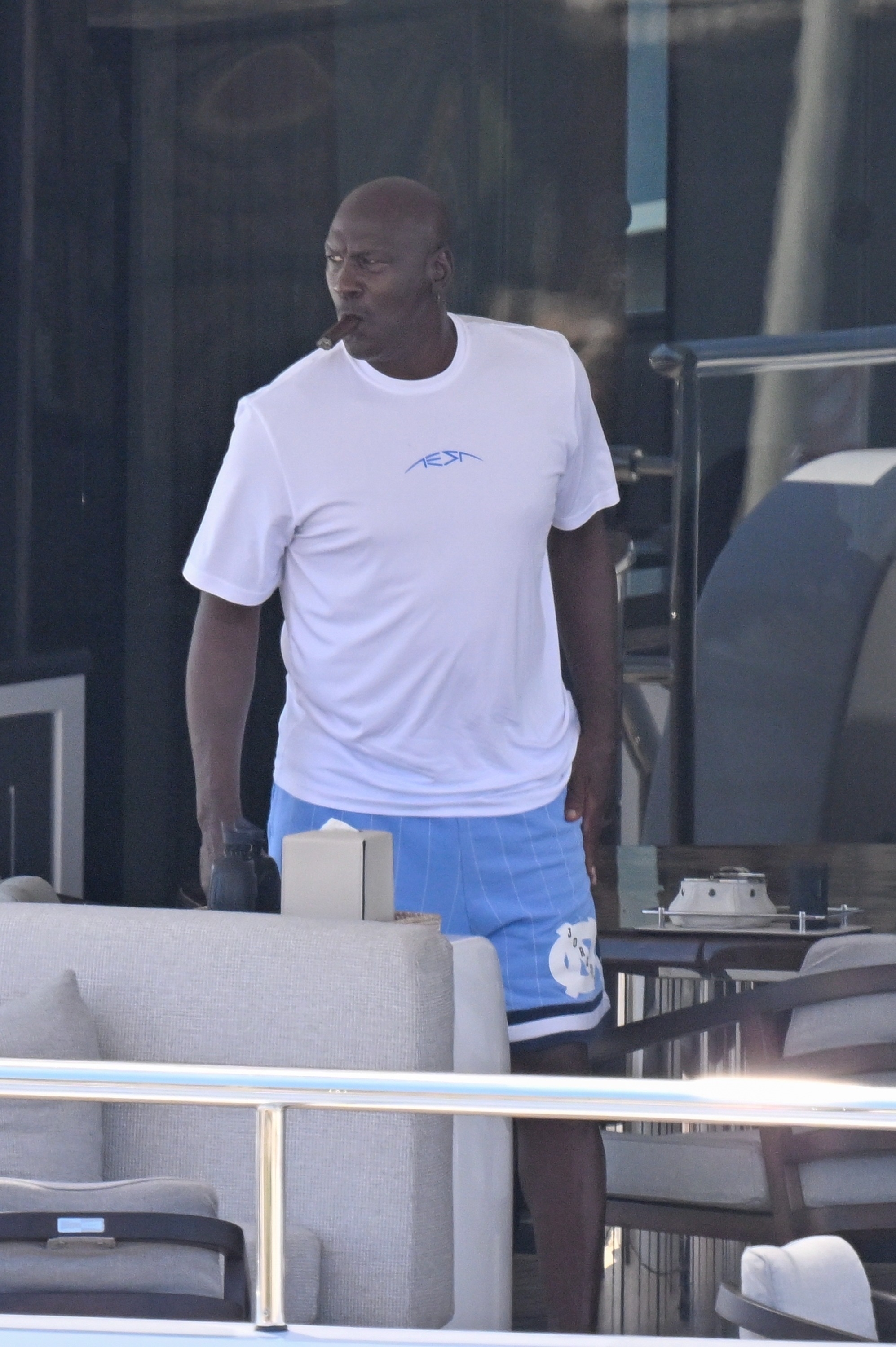 A bald man in a white t-shirt and blue UNC basketball shorts smokes a cigar while standing on the deck of a large yacht.