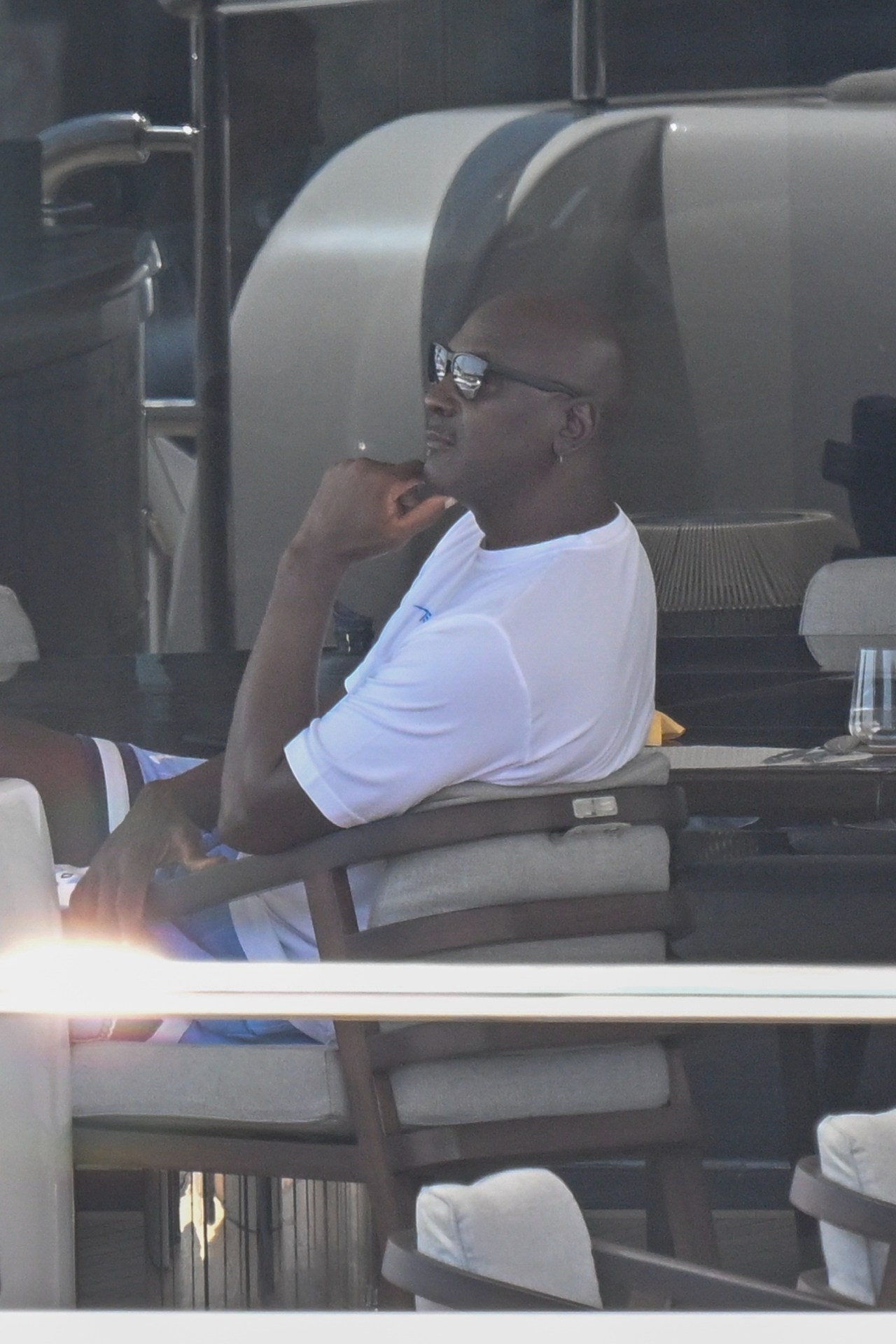 Man in white t-shirt and sunglasses seated on a yacht deck, resting his chin on his fist in a relaxed pose.