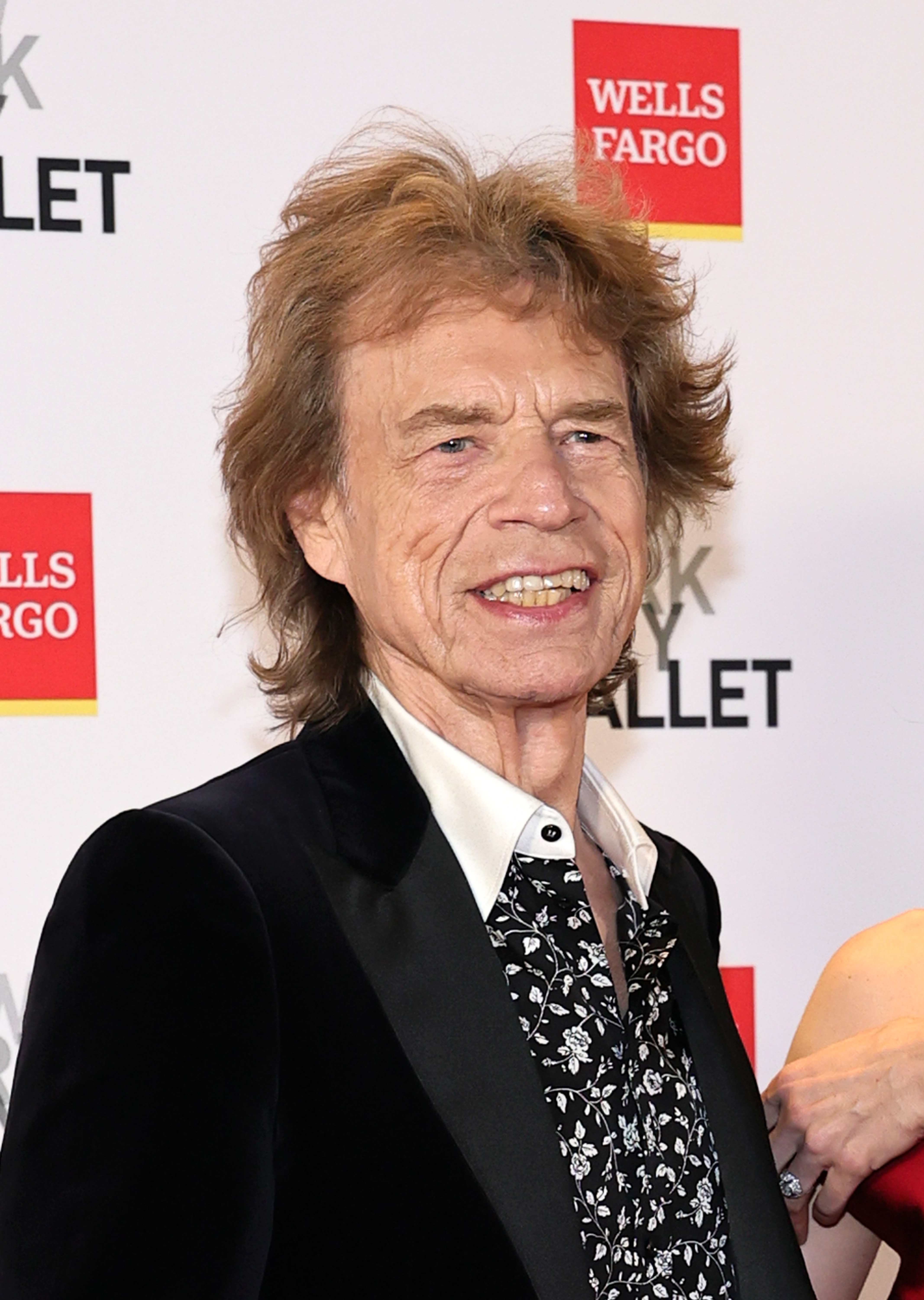 Older man with tousled auburn hair in a black tuxedo jacket and floral shirt smiling at a Wells Fargo and New York City Ballet step-and-repeat backdrop.