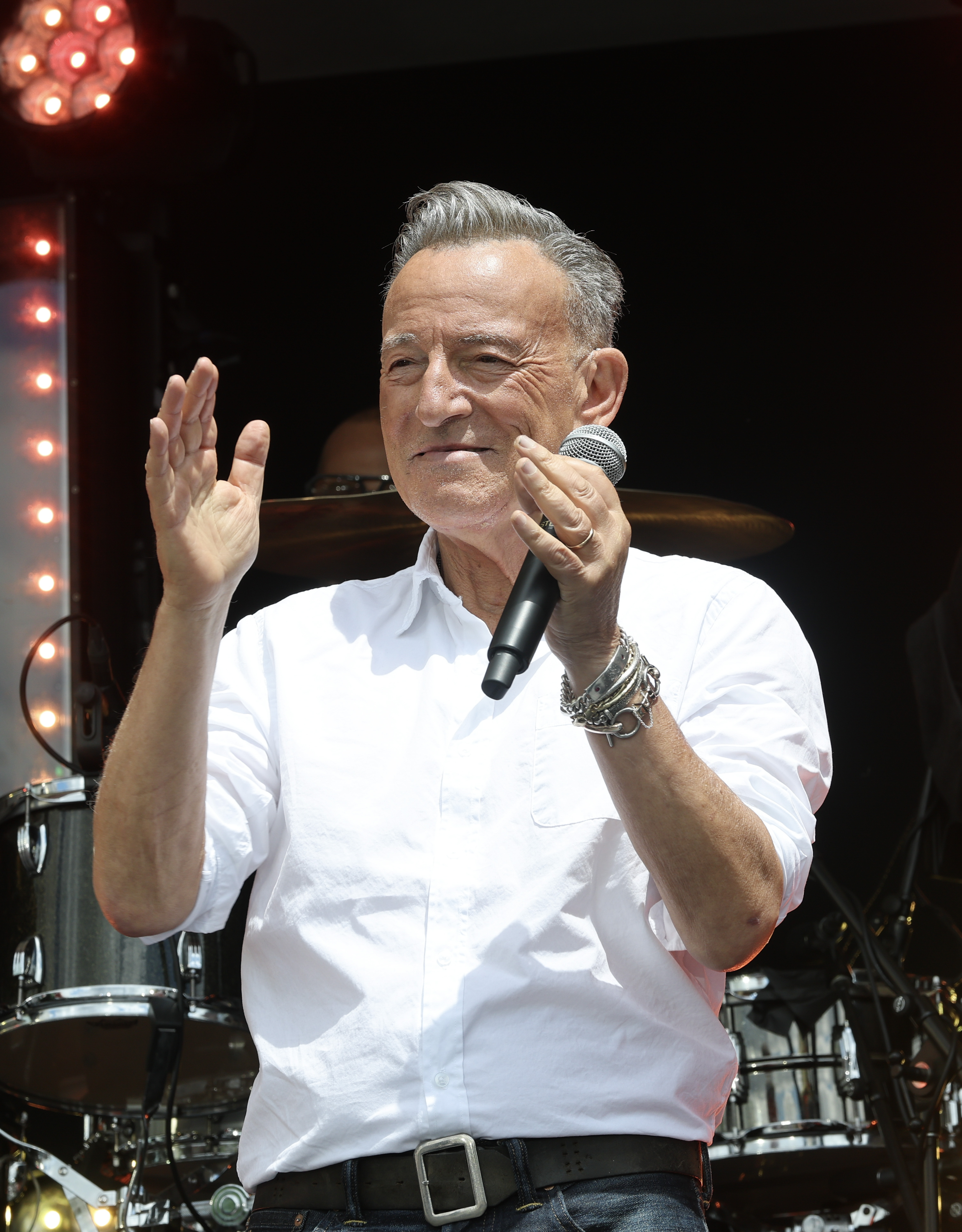An older man with gray hair holds a microphone and raises one hand while performing on an outdoor stage with drum kit visible behind him.