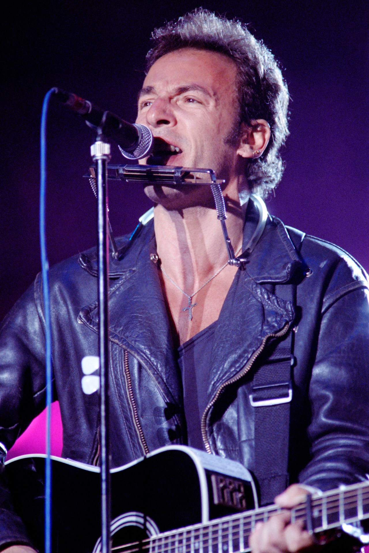 Bruce Springsteen singing at a microphone with a harmonica rack, holding an acoustic guitar, wearing a leather jacket and cross necklace on stage.