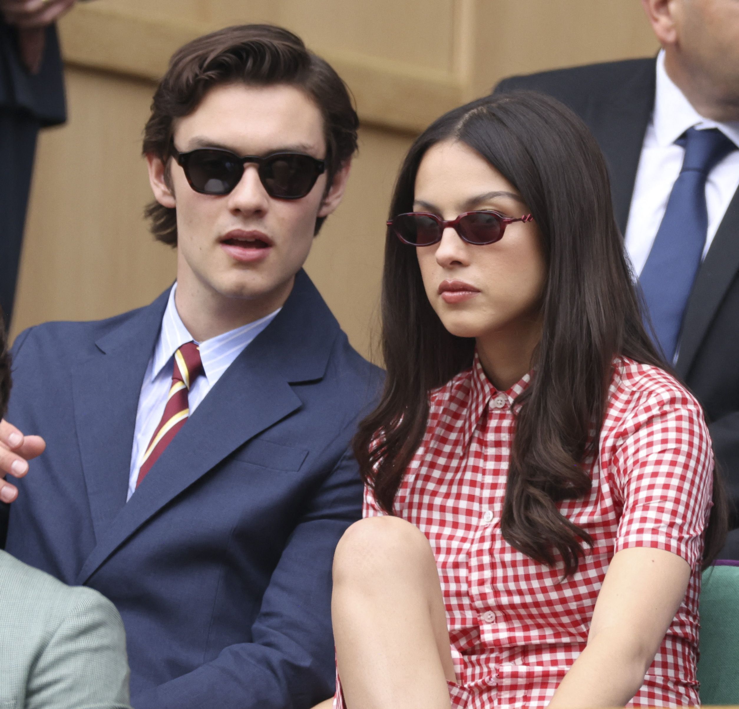 A young man in a navy suit and striped tie with black sunglasses sits beside a young woman in a red gingham dress with small red sunglasses at Wimbledon.