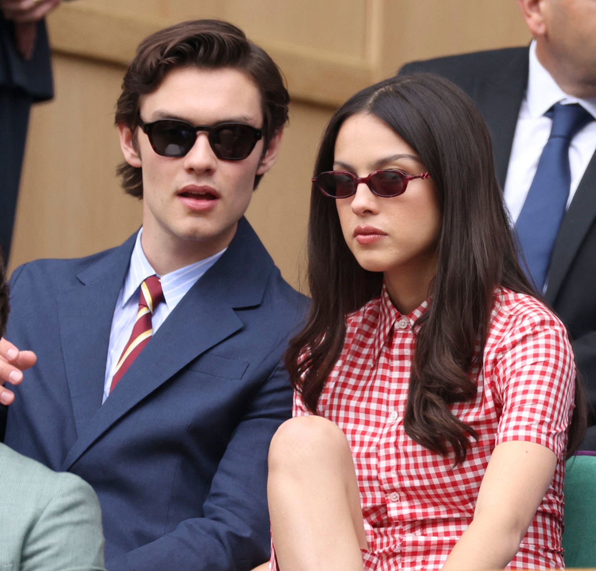 A young man in a navy suit and striped tie with black sunglasses sits beside a young woman in a red gingham dress with small red sunglasses at Wimbledon.