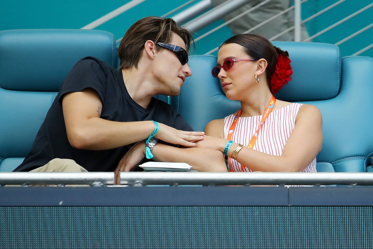 A young man in a black tee and sunglasses leans toward a young woman in a red-striped top with a red flower in her hair, seated at a tennis stadium.
