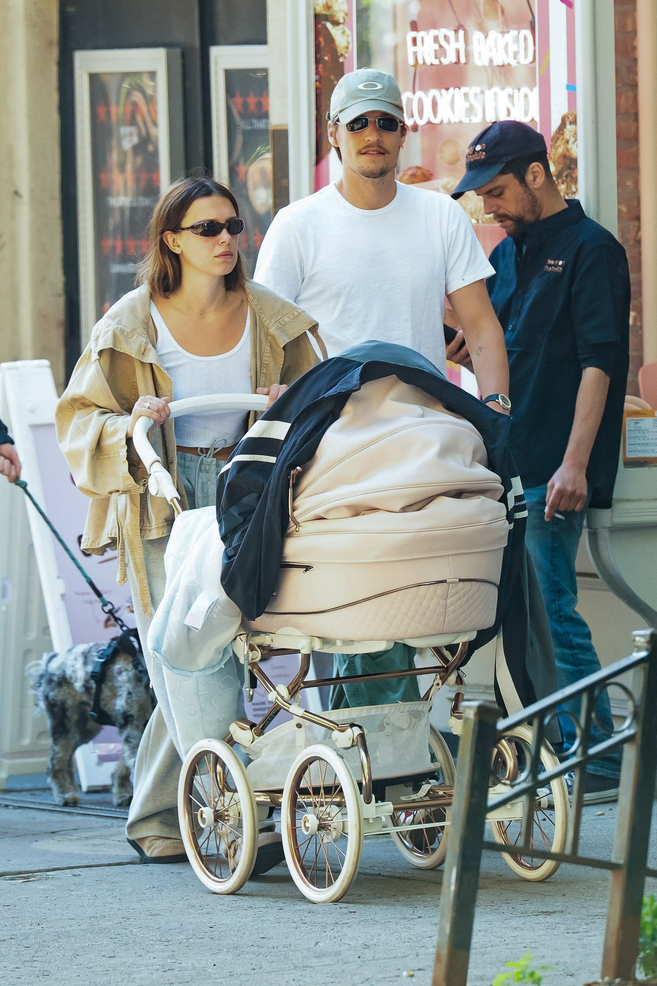 A woman in gray jeans and a tan jacket pushes an elegant blush stroller on a city sidewalk while a man in a white tee and cap walks alongside her.