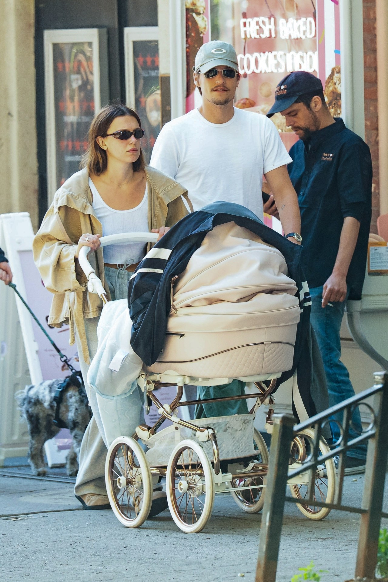 A woman in gray jeans and a tan jacket pushes an elegant blush stroller on a city sidewalk while a man in a white tee and cap walks alongside her.