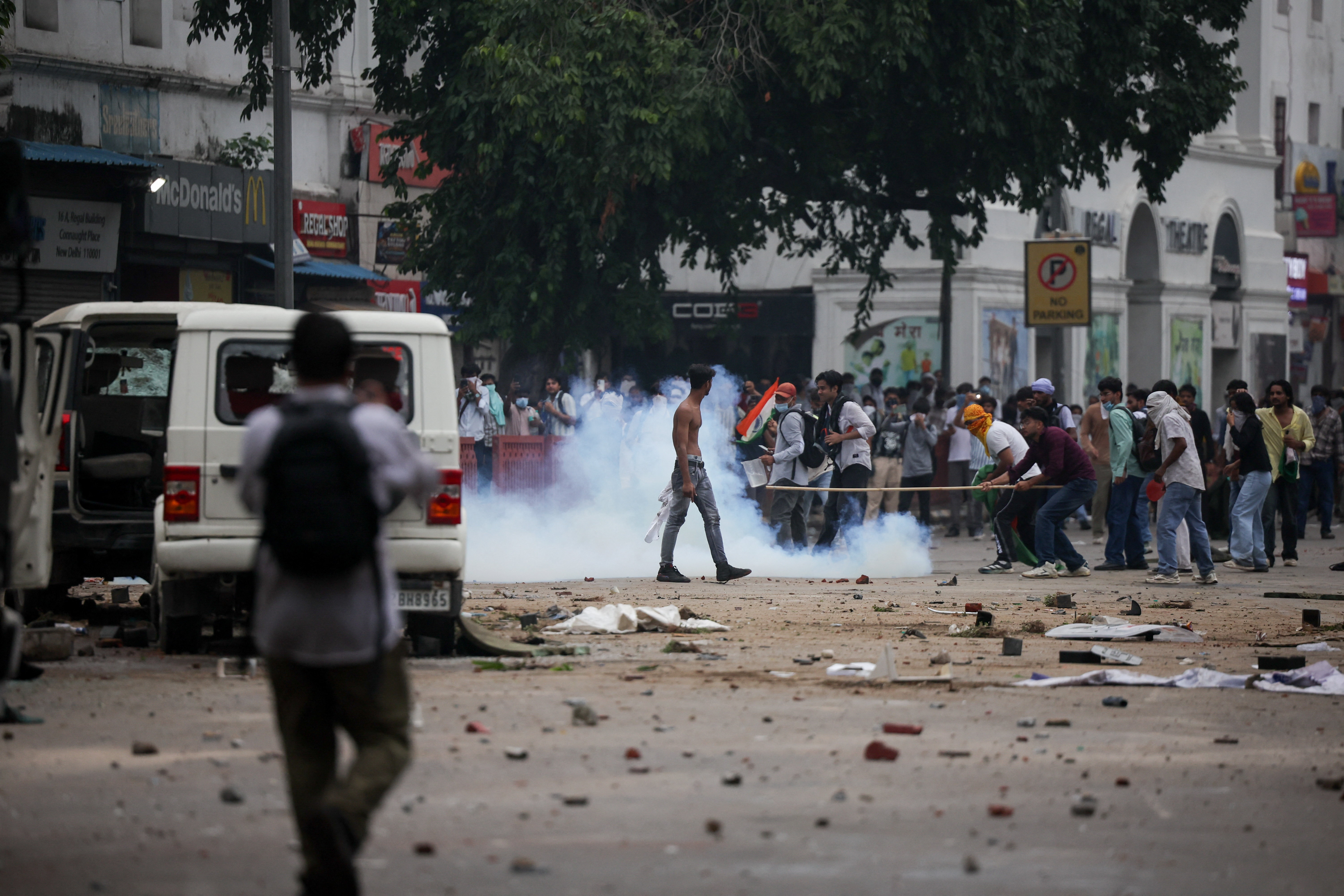 A wide street strewn with debris as tear gas smoke engulfs protesters near a McDonald's and a Regal Theatre sign in New Delhi.