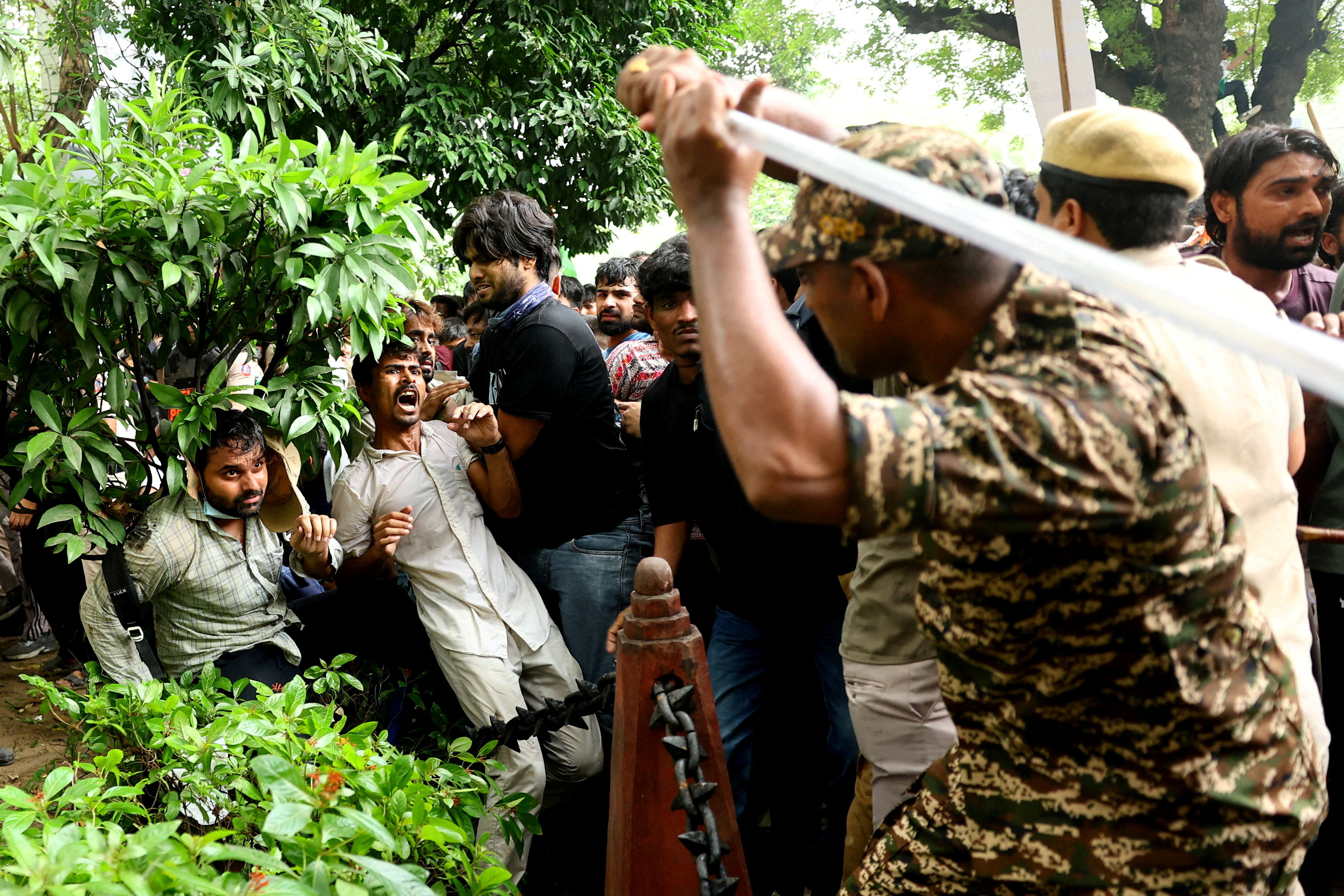 A uniformed officer in camouflage raises a baton over a crowd of protesters who are pressed against barbed wire fencing amid trees.