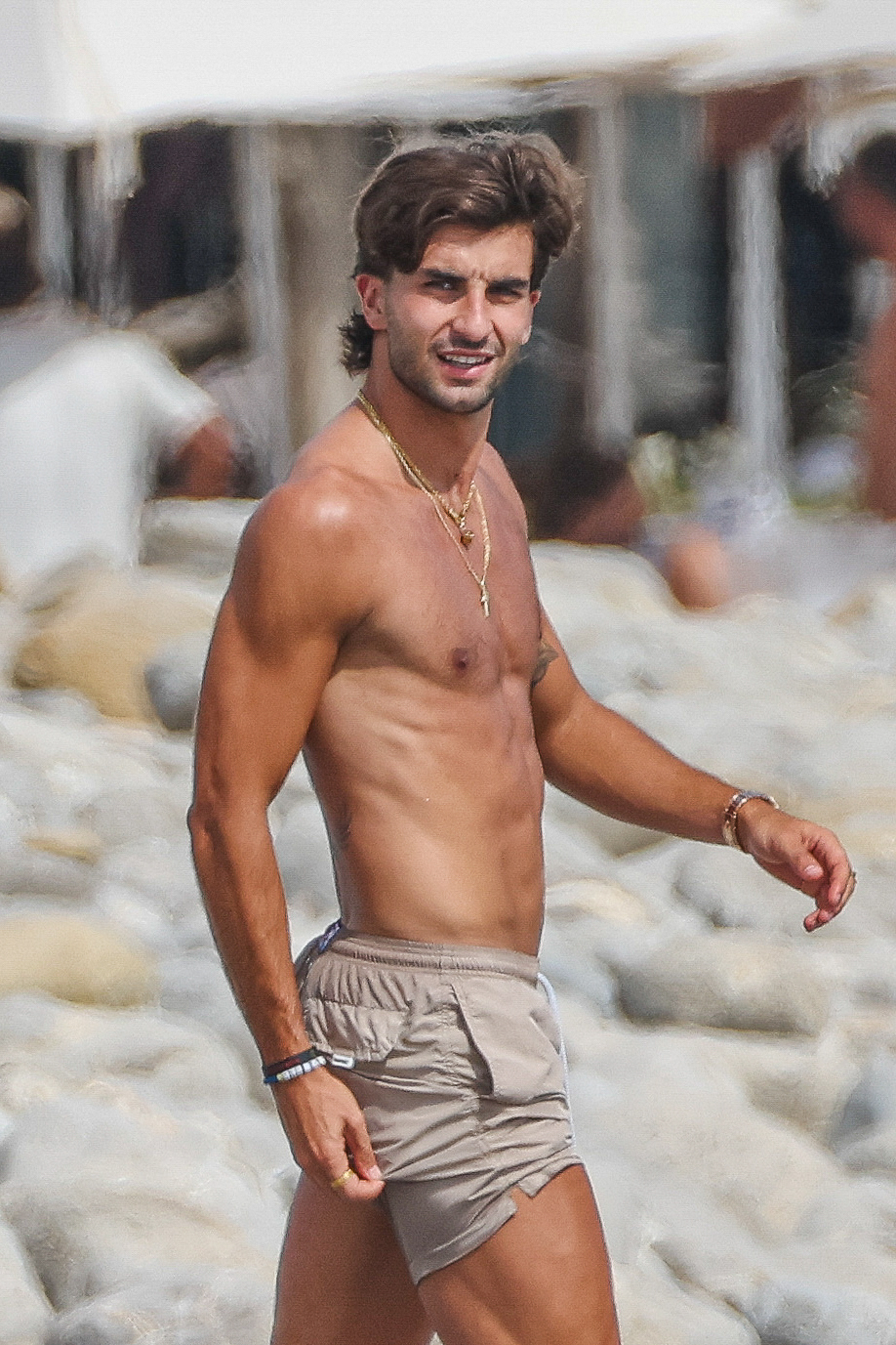 Click to open the image gallery Shirtless young man with dark curly hair and gold necklaces smiling on a rocky beach.