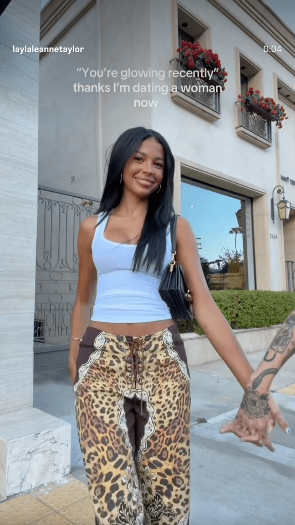 Woman in white crop top and leopard-print pants smiling outdoors, holding hands with a tattooed person, with text overlay about dating a woman.