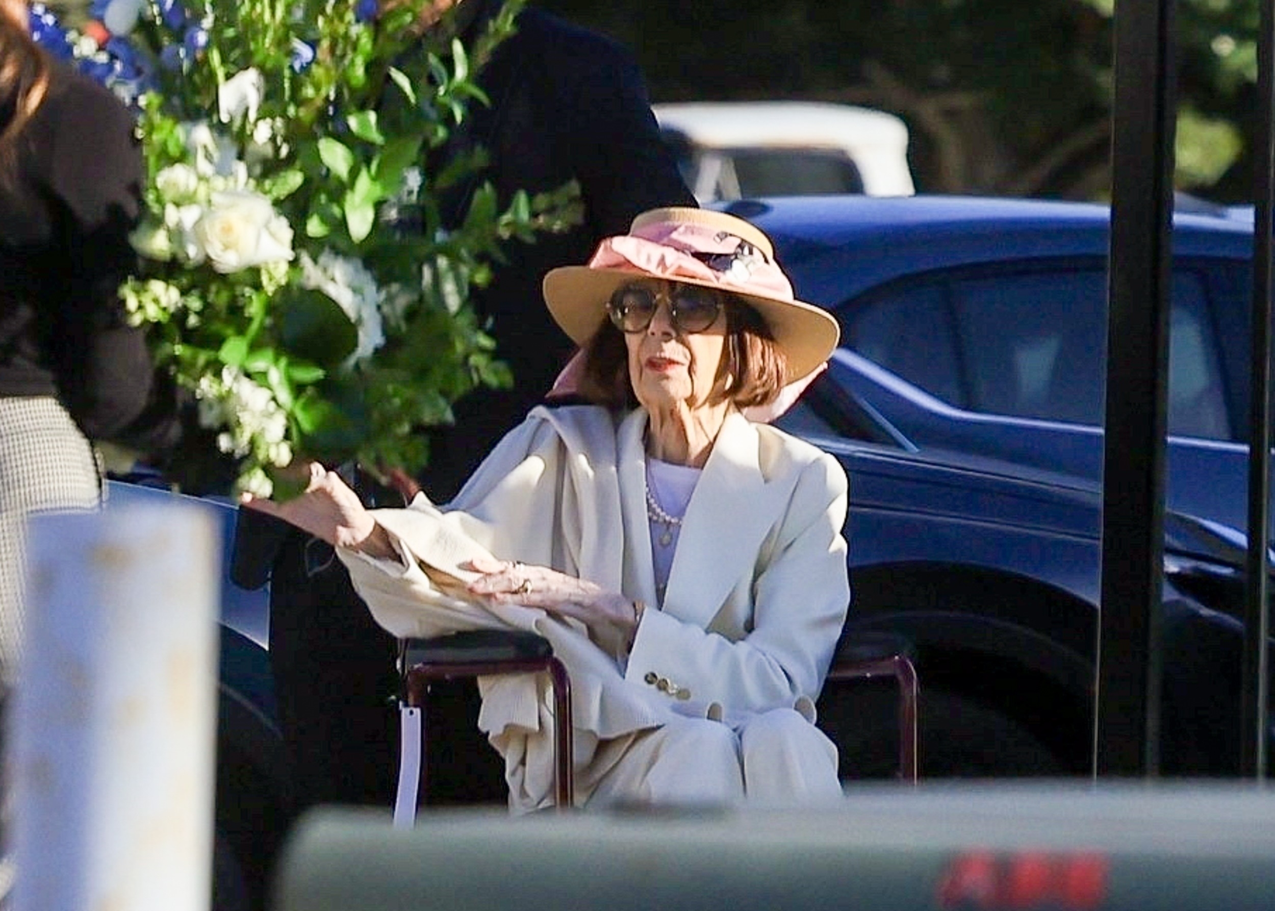 Elderly woman in a cream coat and wide-brimmed tan and pink hat seated in a wheelchair outdoors near a floral arrangement.