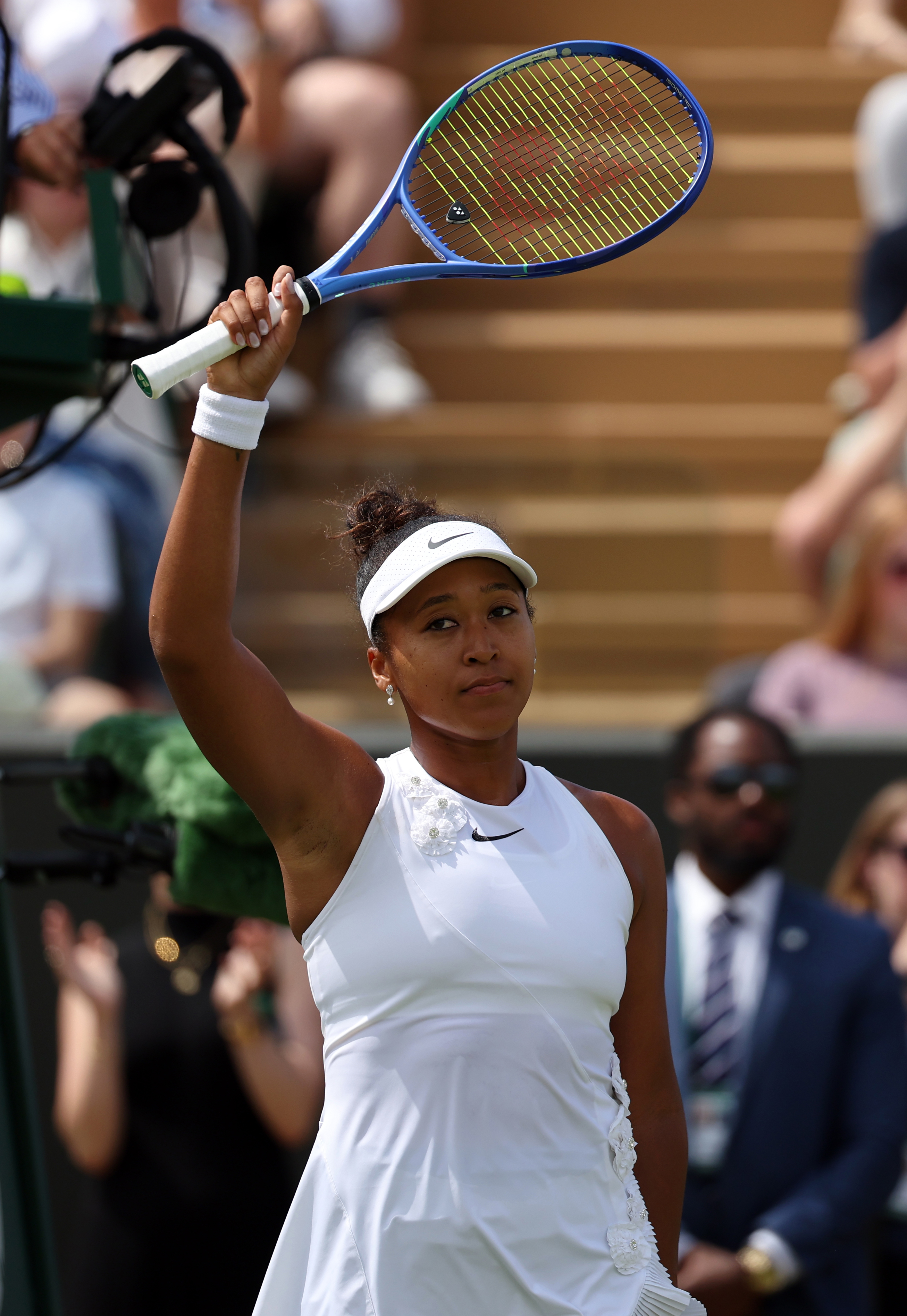 Click to open the image gallery Naomi Osaka in a white Nike dress and visor raises her blue Wilson racket overhead toward the crowd at Wimbledon.