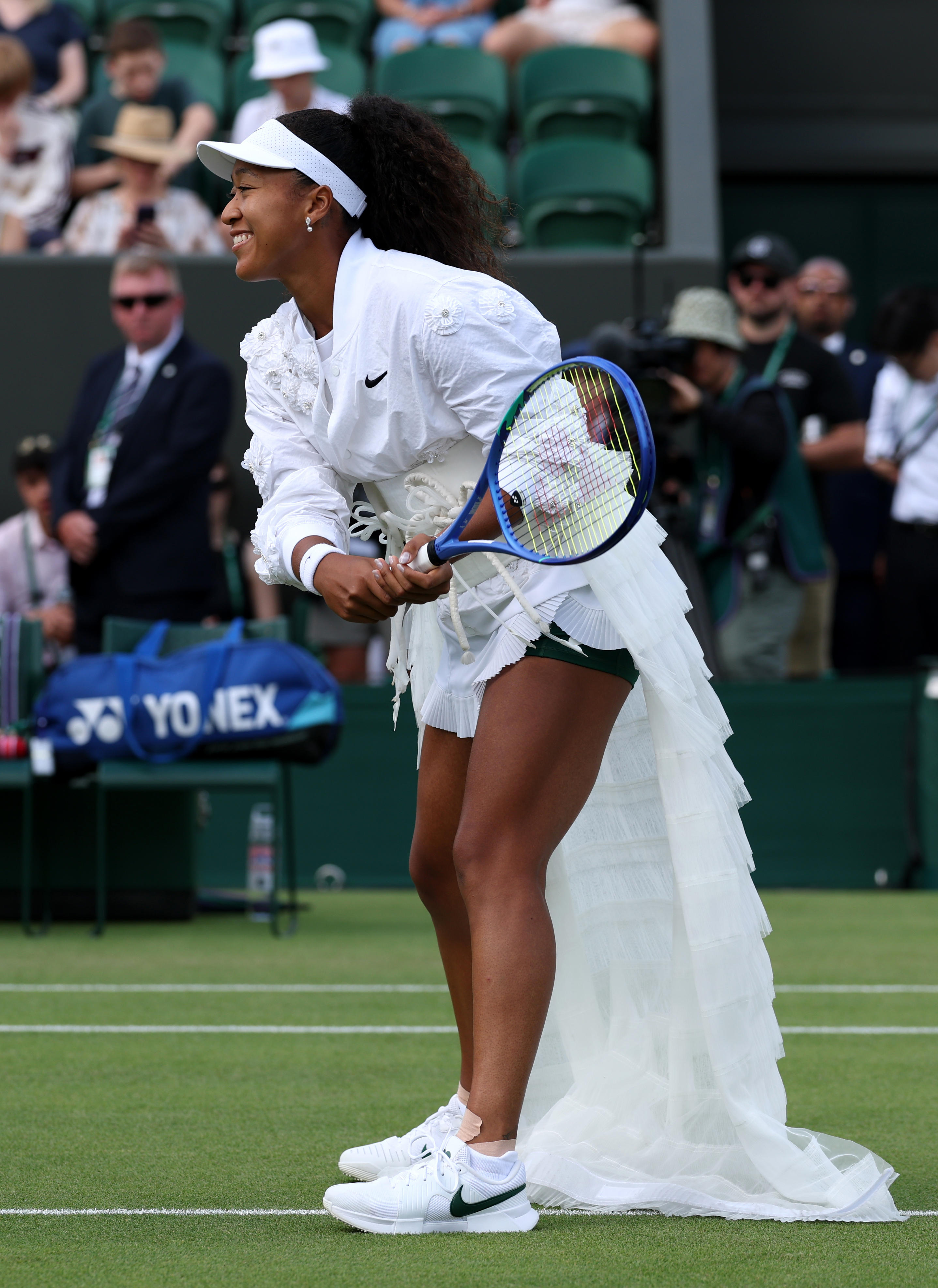 Click to open the image gallery Naomi Osaka smiling and bent forward on a grass court in a white floral ruffled outfit, holding a blue tennis racket.
