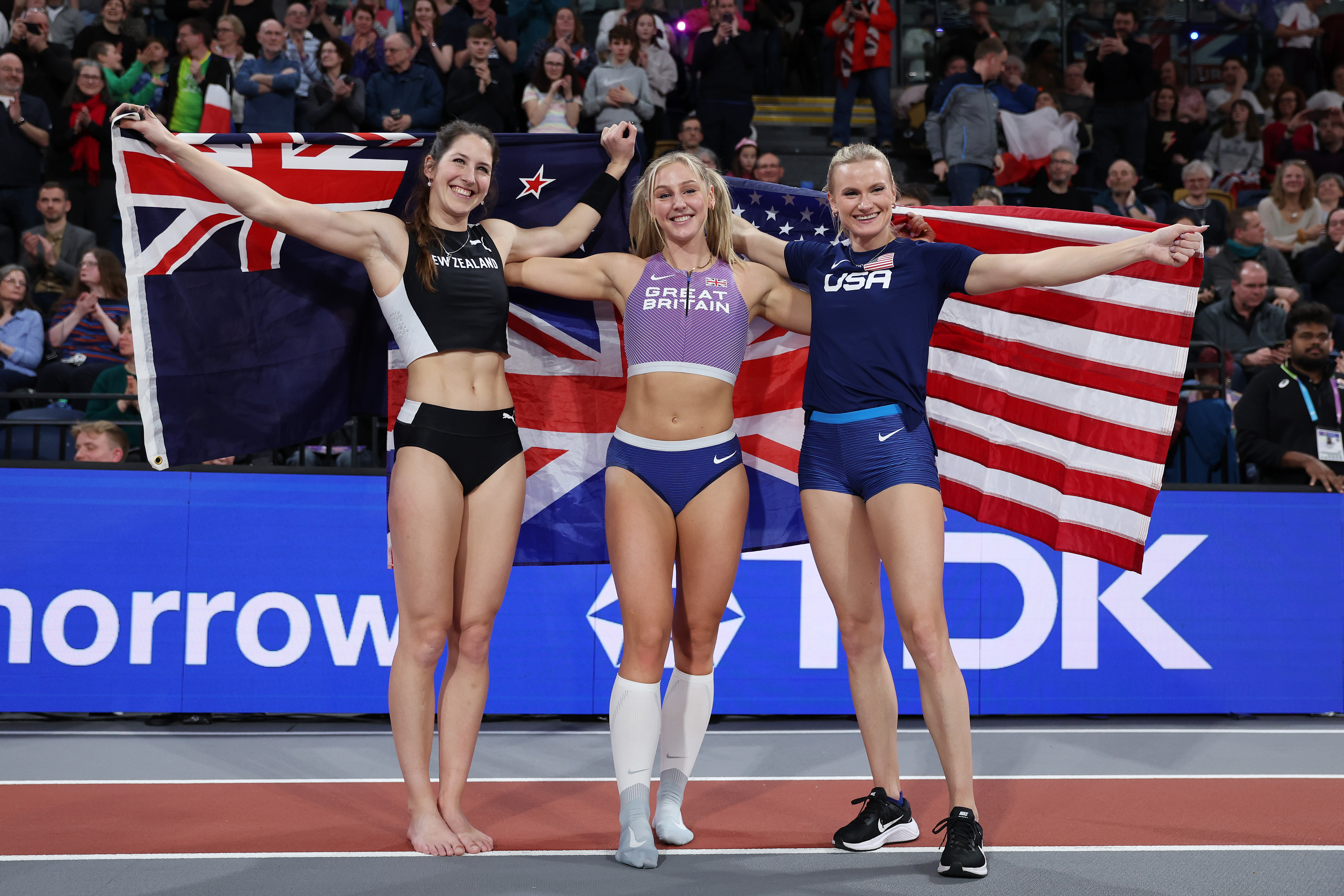 Click to open the image gallery Three female pole vaulters hold up the New Zealand, Great Britain, and US flags while smiling at an indoor athletics arena.