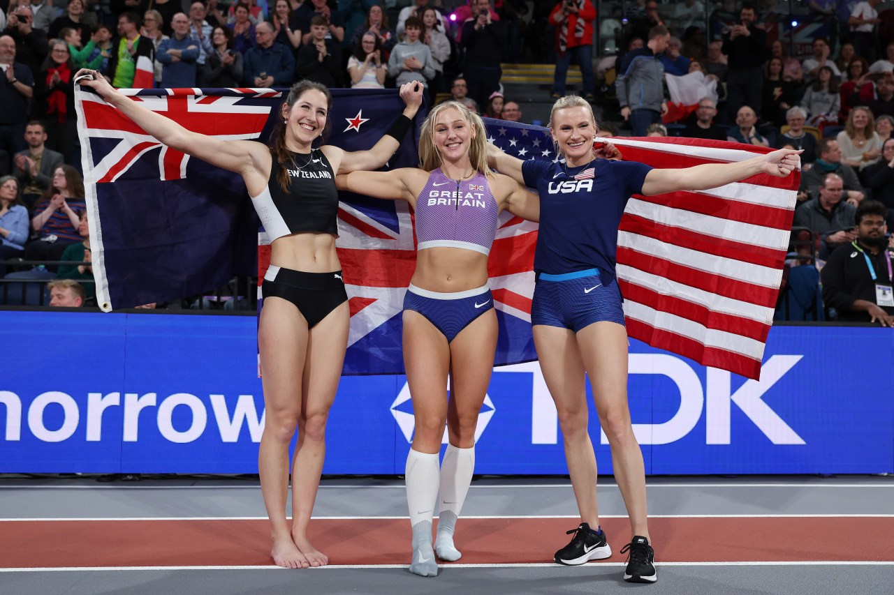 Three female pole vaulters hold up the New Zealand, Great Britain, and US flags while smiling at an indoor athletics arena.