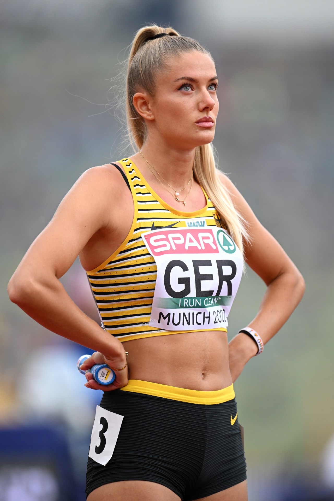 A German female sprinter in a striped yellow and black crop top holds a relay baton at her side on the track, looking off into the distance.