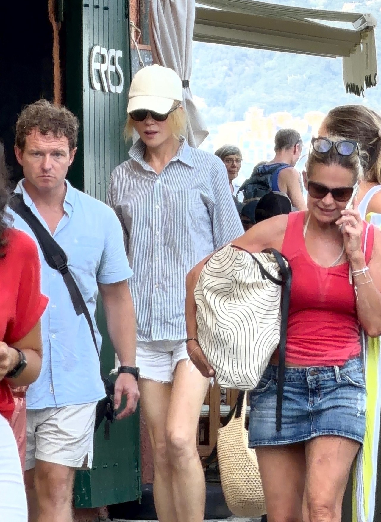 Woman in striped shirt, white shorts, and cream baseball cap walks through a crowd near a shop in Portofino.