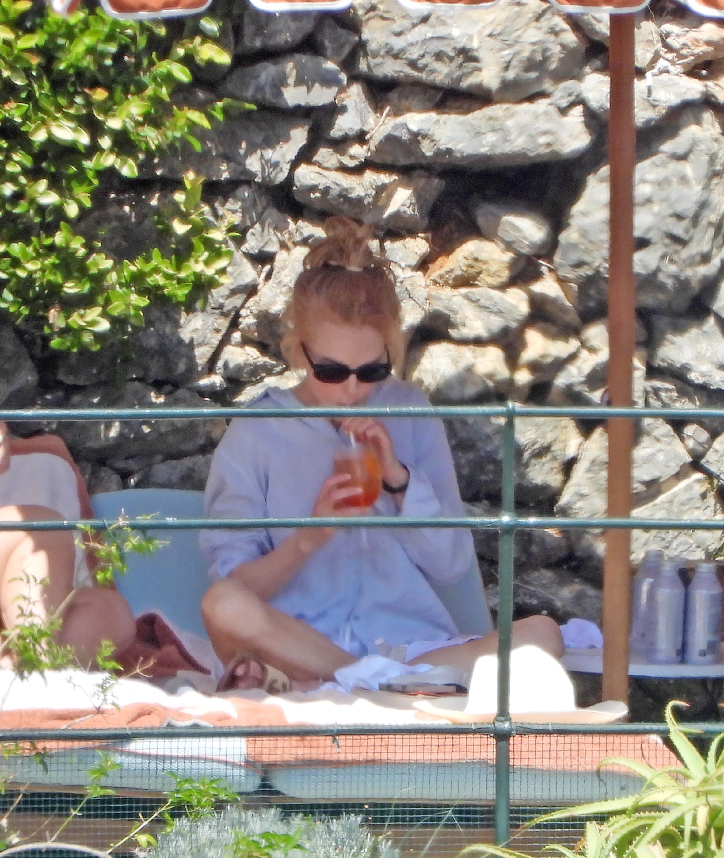 Woman with blonde updo and black sunglasses sitting on a sun lounger, sipping an orange drink through a straw, with a stone wall behind her.