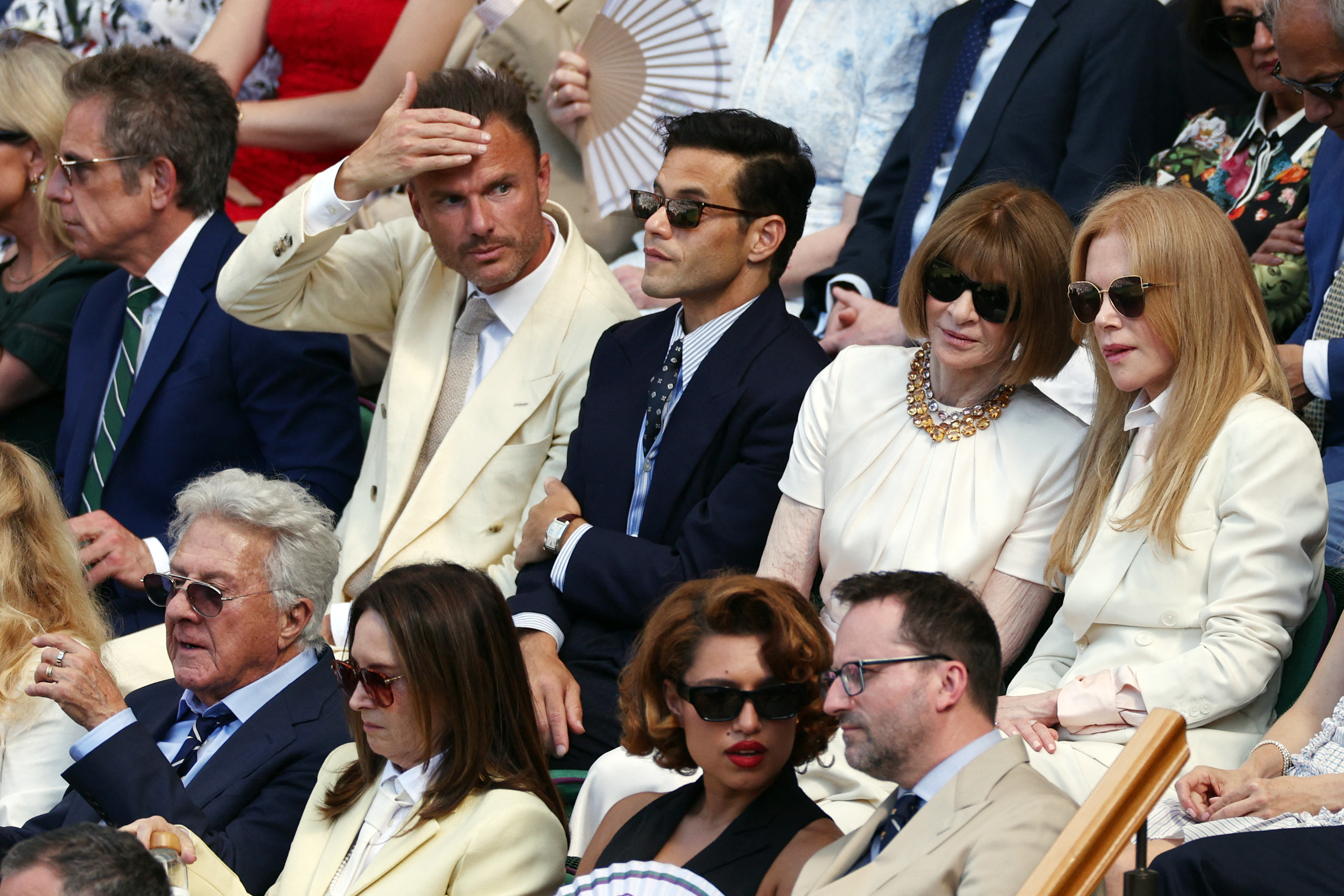A group of celebrities seated in Wimbledon's Royal Box, including a man in a navy suit with sunglasses, a woman in white with a statement necklace, and a blonde woman in cream.