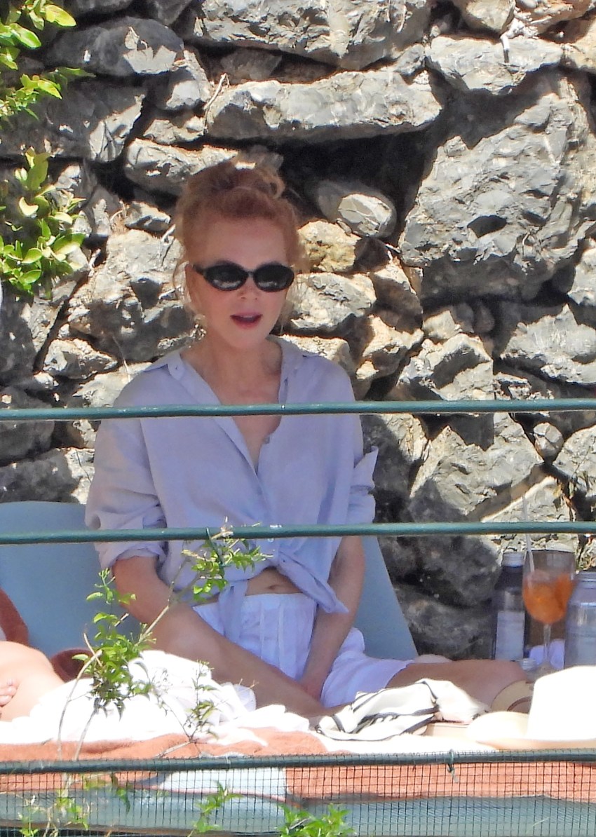 Woman with auburn updo and black oval sunglasses sitting poolside in a pale blue shirt and shorts, with Aperol Spritzes on the table, rocky cliff backdrop behind her.