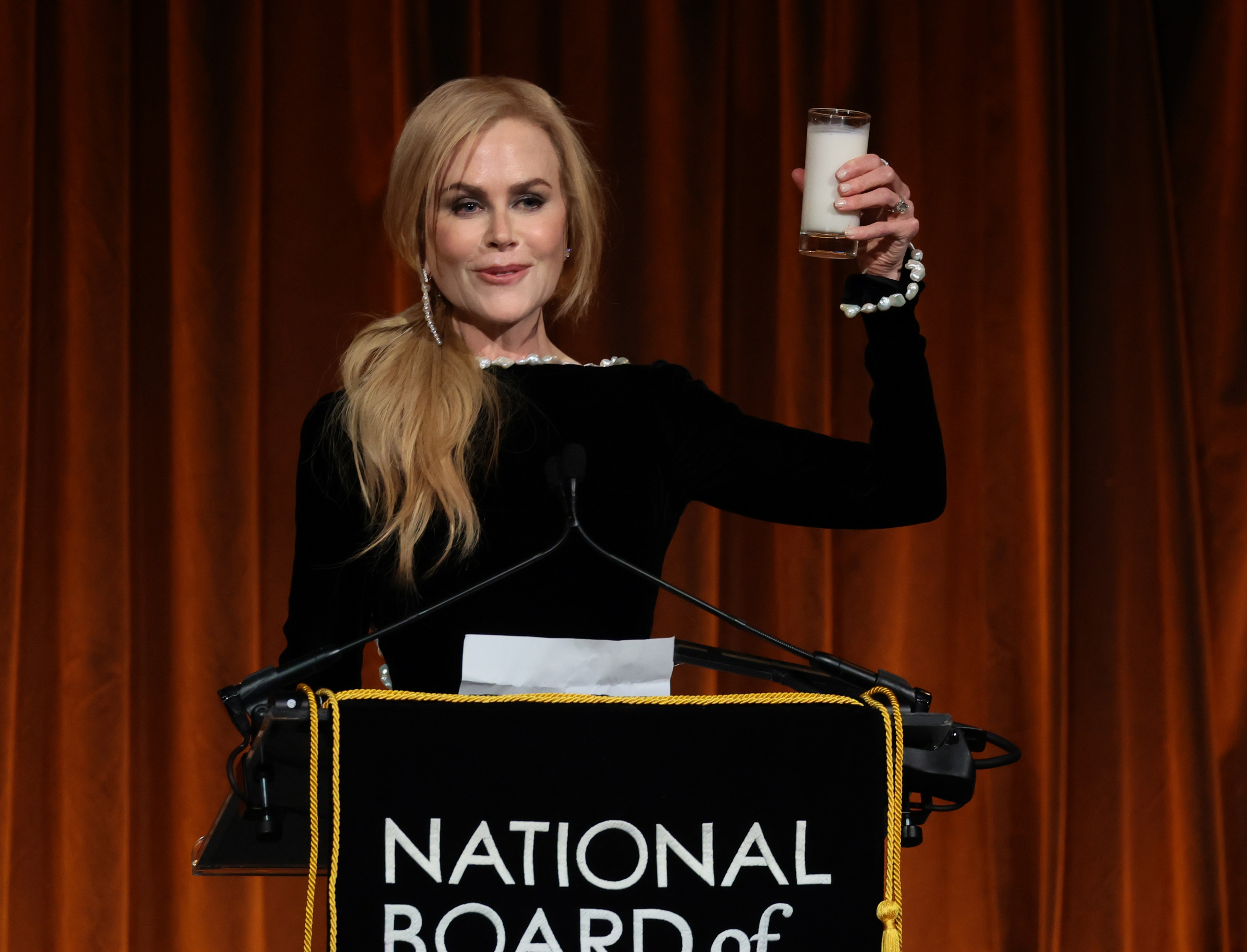 Woman in black long-sleeve dress with pearl trim raises a glass at a podium bearing the National Board of Review logo.