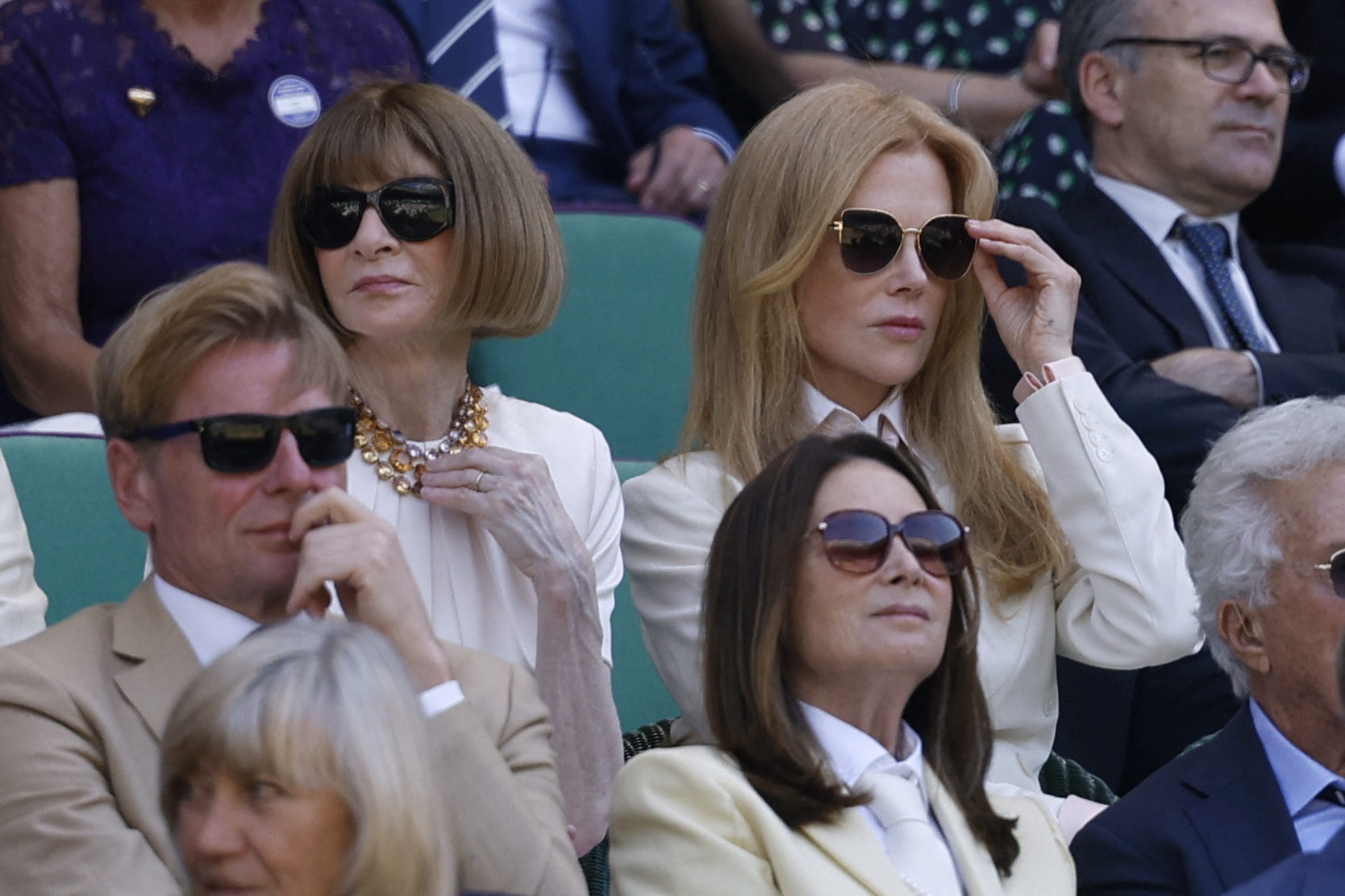 Two women wearing sunglasses seated in the Wimbledon Royal Box, surrounded by other spectators in green chairs.