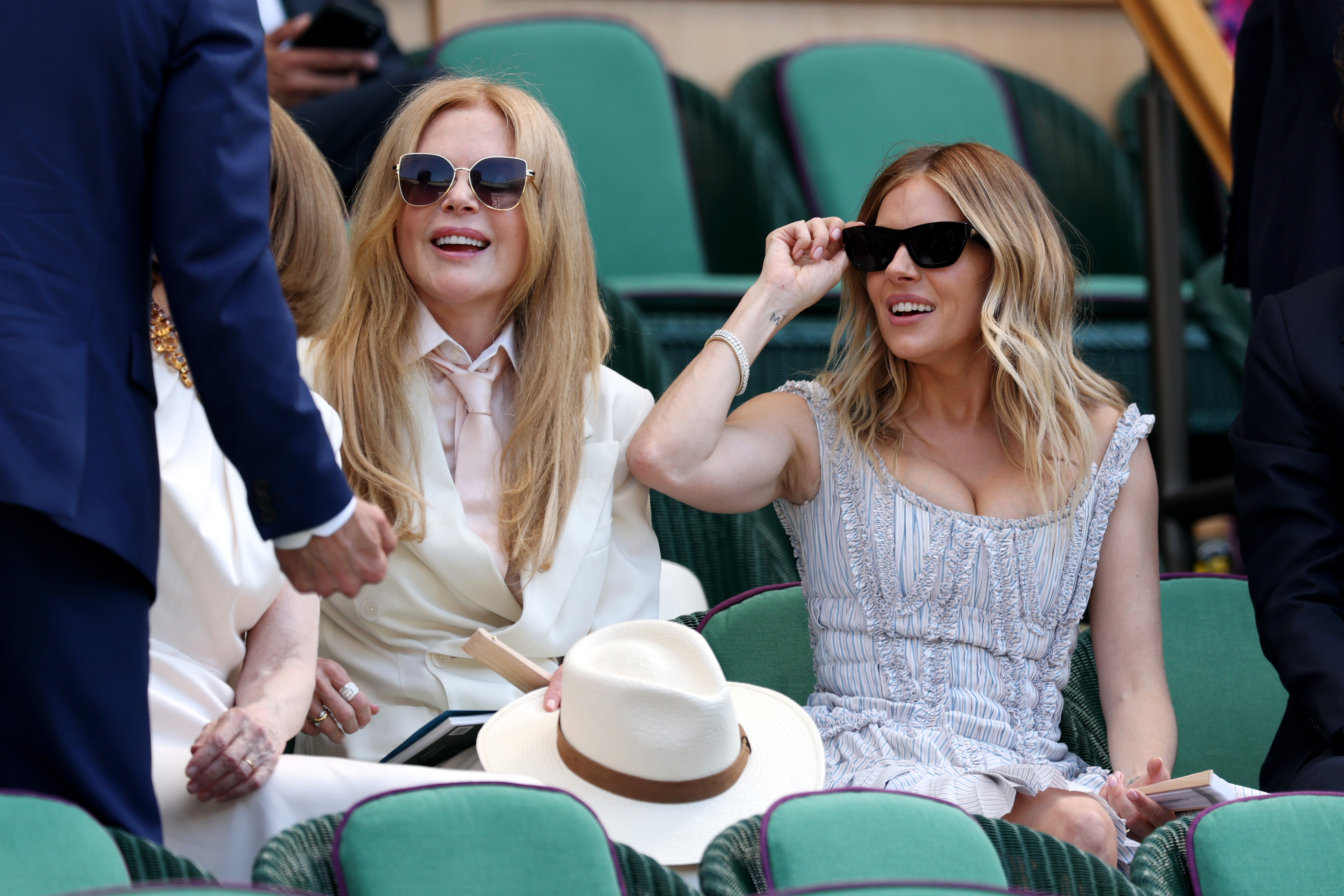 Two smiling women in sunglasses seated side by side in green Royal Box chairs at Wimbledon, one in a cream suit and one in a light blue dress.