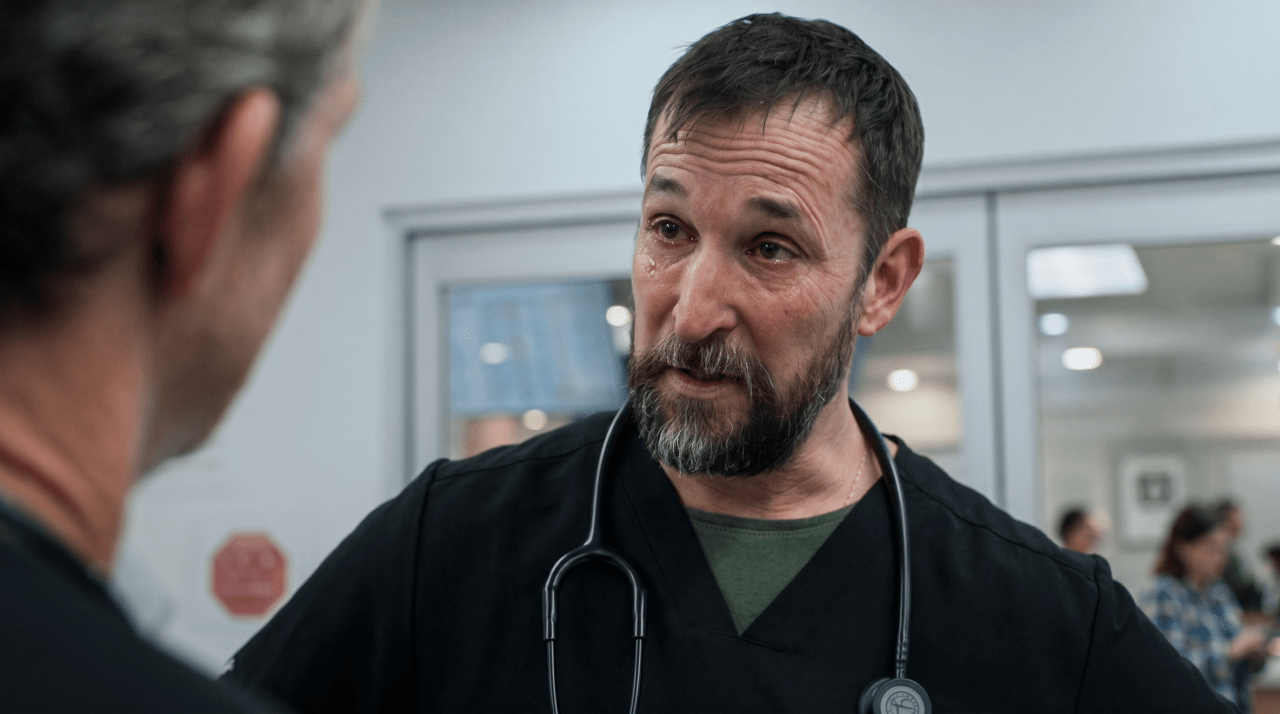 Bearded man in black scrubs with stethoscope talking to another person in a hospital hallway.