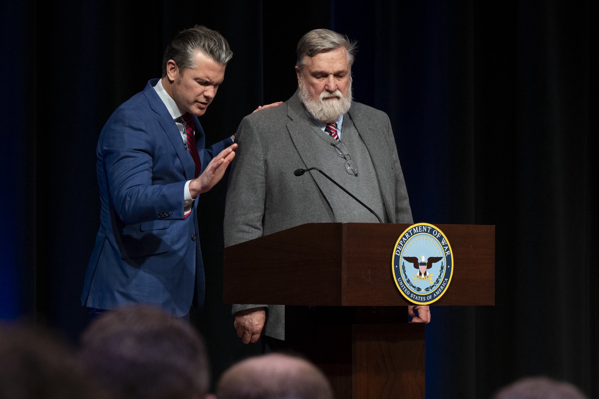 Two men in suits stand at a Department of War podium; the younger man in a blue suit places his hand on the older bearded man's shoulder.