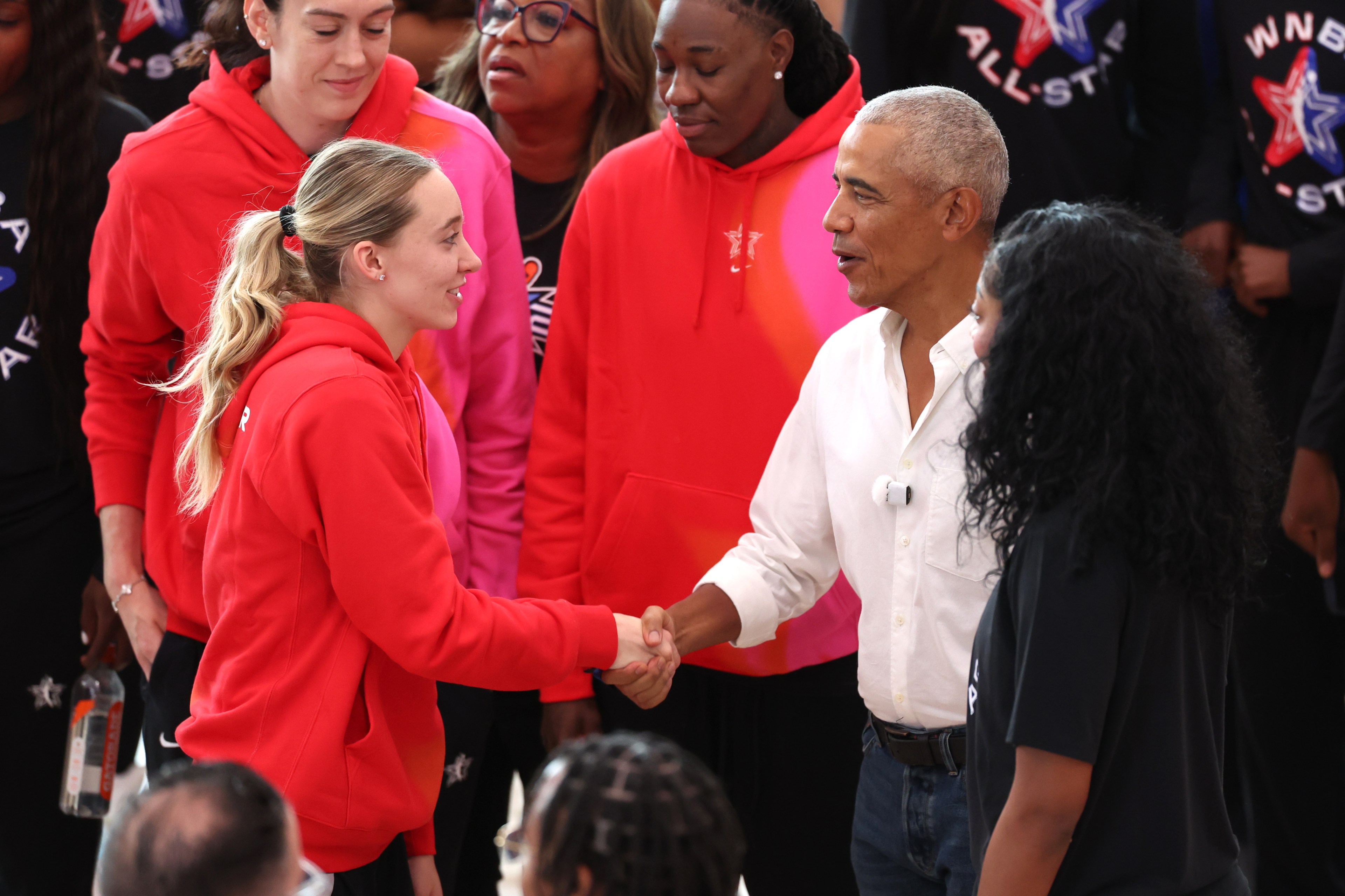 Barack Obama shaking hands with Paige Bueckers, surrounded by WNBA players in red All-Star hoodies, at an indoor practice facility.