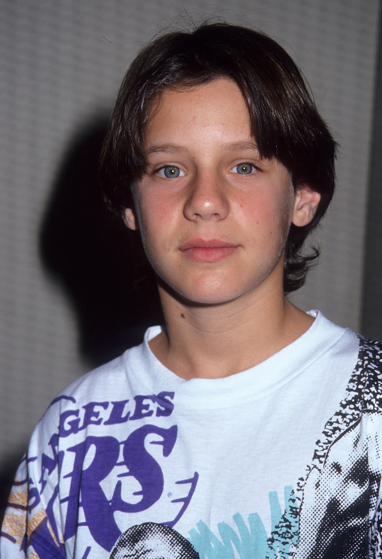 Young dark-haired boy in a white Los Angeles graphic t-shirt posing against a gray wall at a press event.