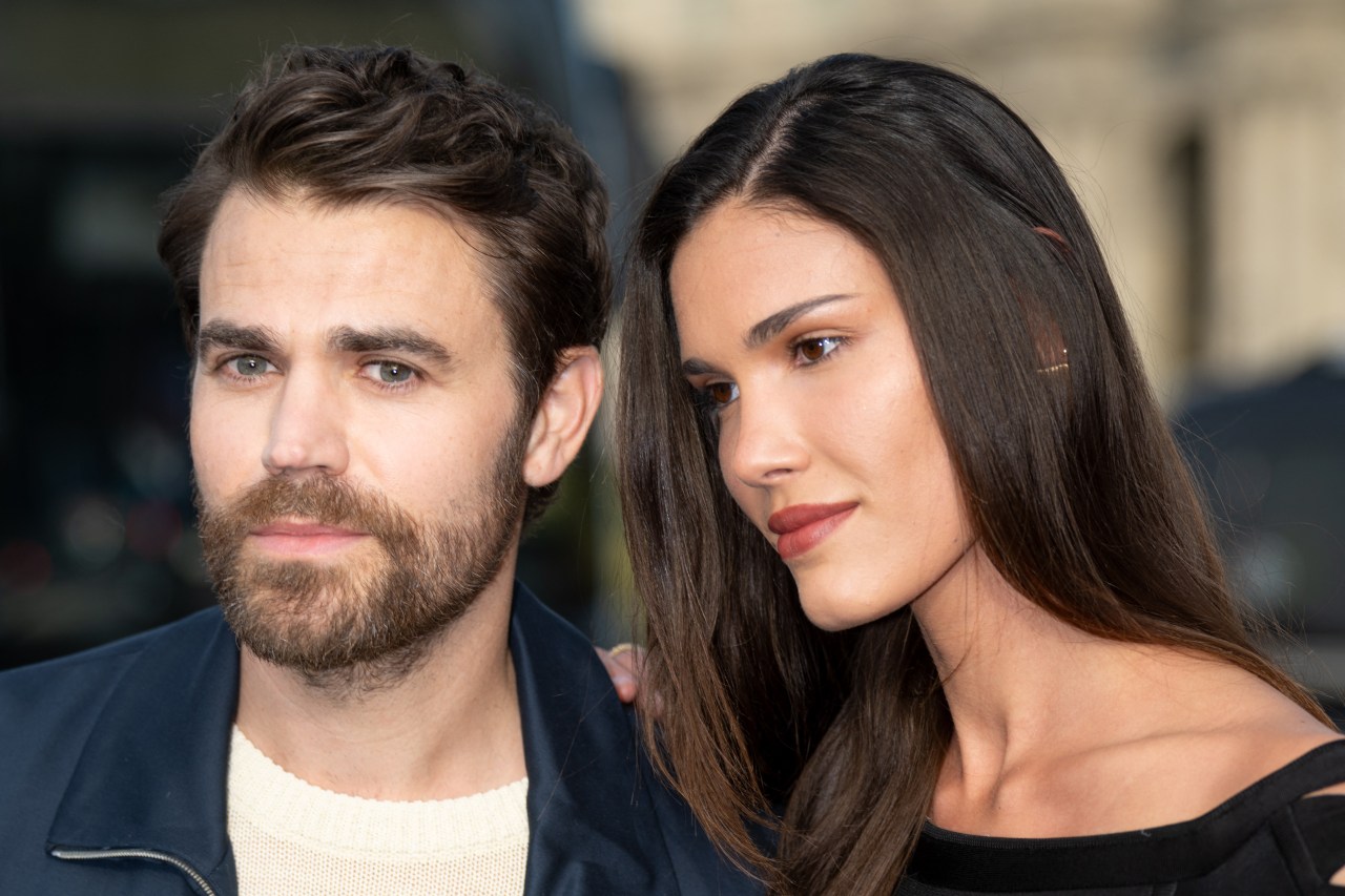 A bearded man in a navy jacket and cream sweater stands close to a woman with long dark hair in a black off-shoulder top.