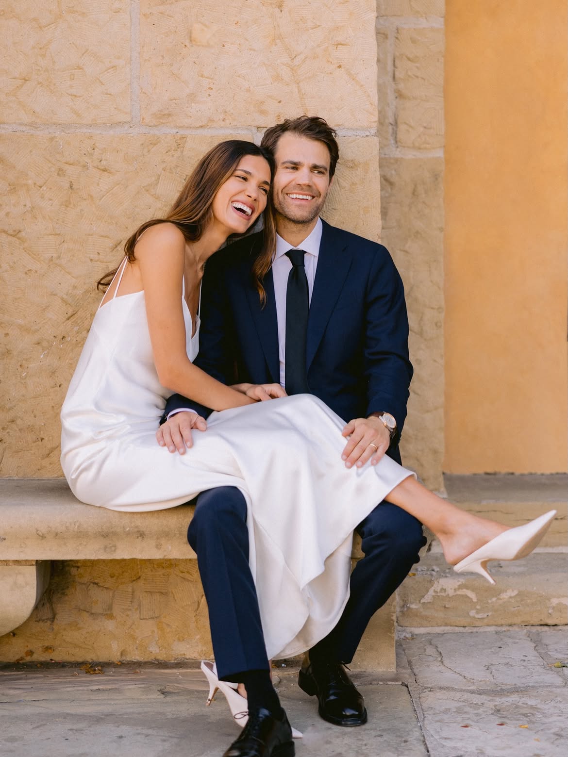 Bride in a white slip dress sits on groom's lap as they both laugh, seated against a stone wall outdoors.