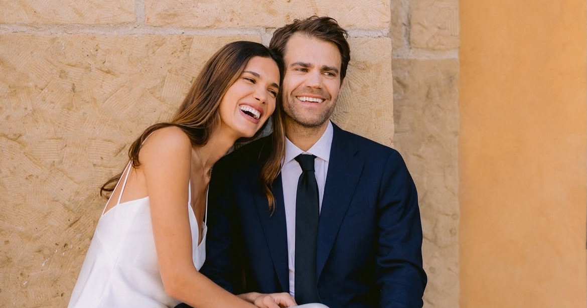 A bride in a white slip dress and groom in a navy suit sit laughing together on a stone bench against a warm yellow wall.