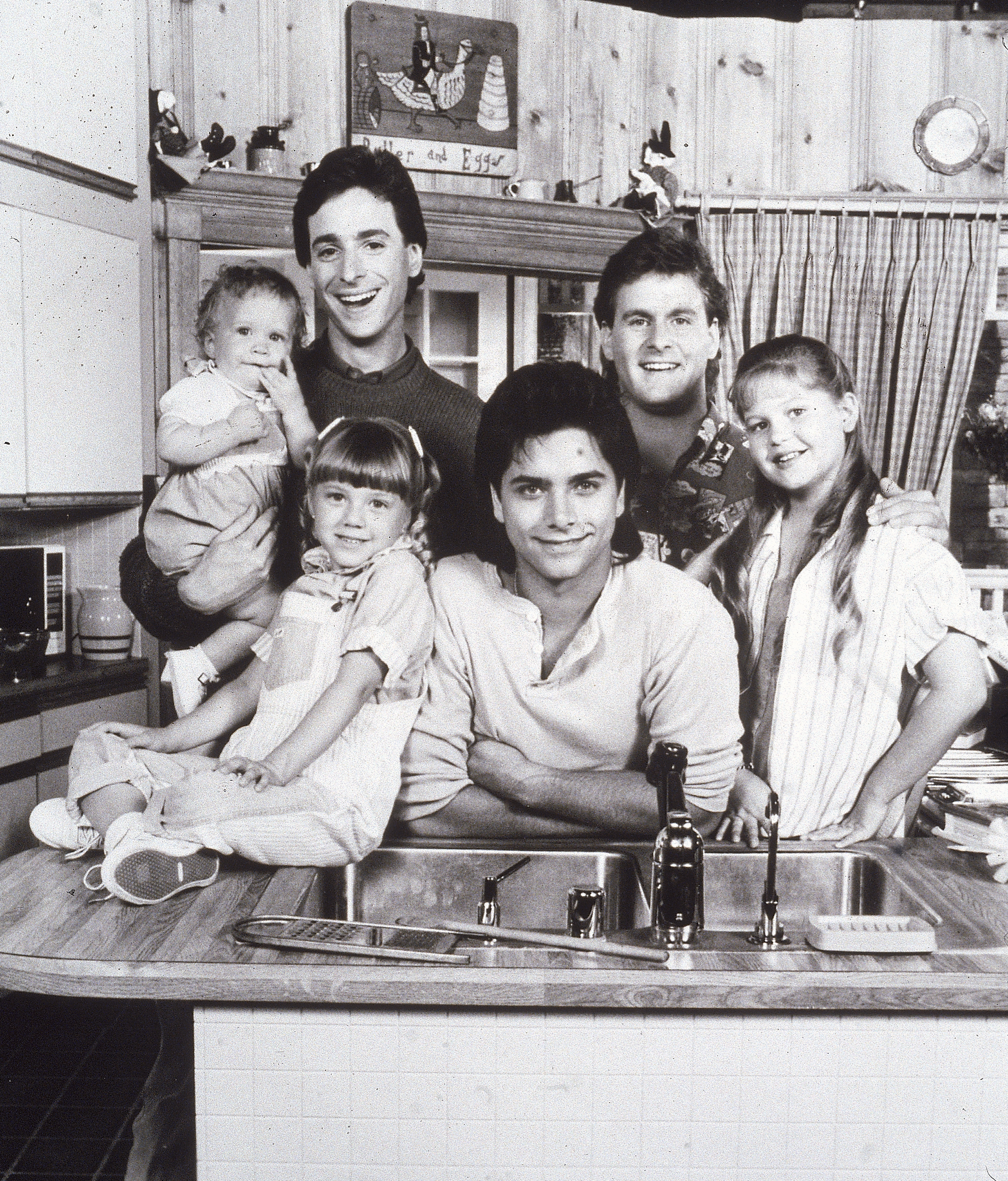 Black-and-white promo photo of the Full House cast posing around a kitchen sink, including two adult men, one young man, two girls, and a baby, circa 1987.